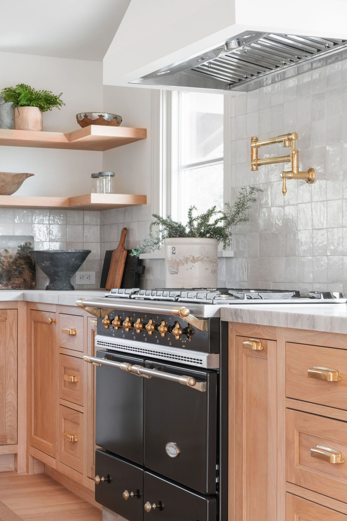 Kitchen with wooden cabinets, open shelves, a stove, and a window with plants, white tiled backsplash, and brass faucet.