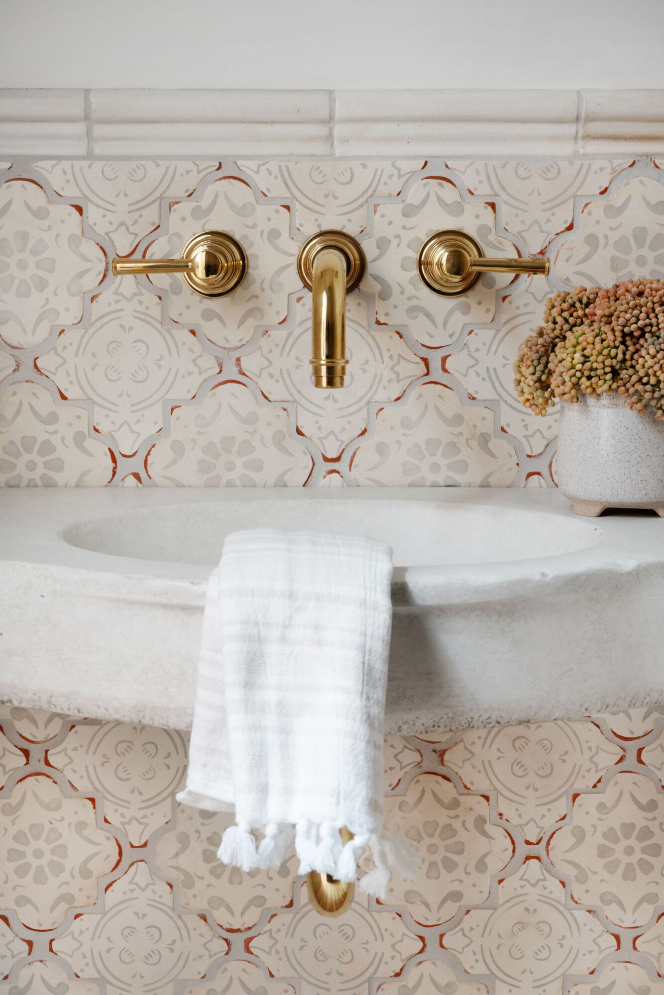 Bathroom sink with gold faucet and handles, white towel hanging, patterned tile background, and a potted plant.