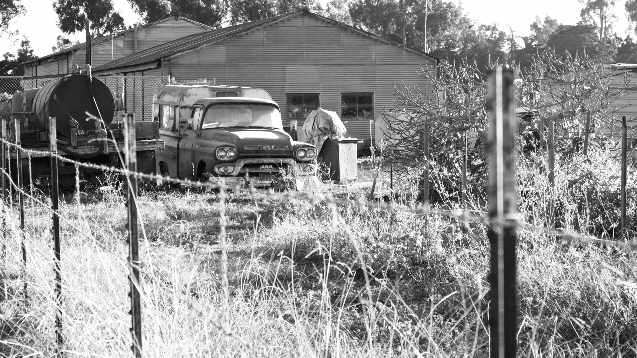 A black and white photo of an outdoor scene with a fence, overgrown grass, and a building with vehicles parked nearby.