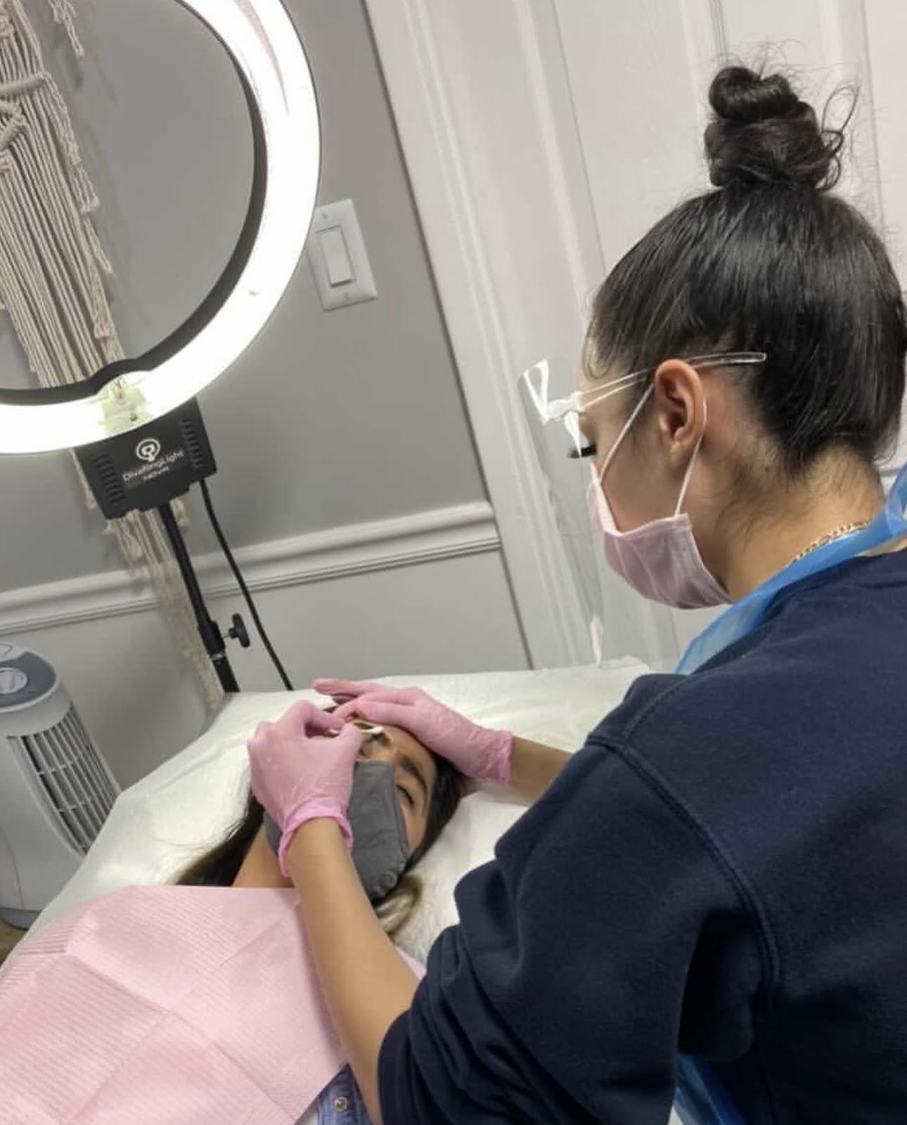 A woman wearing glasses, a pink mask, and pink gloves performs a facial treatment on a person lying down, in a clinical setting.