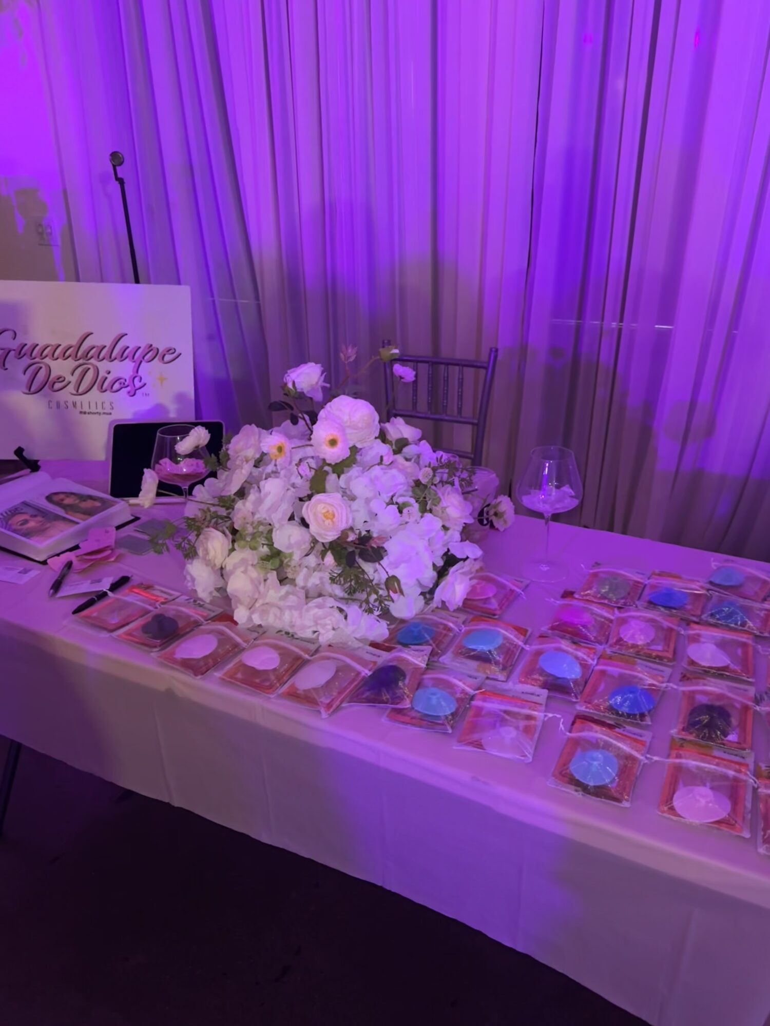 Table with floral centerpiece, makeup products, and a sign, set against purple curtains, under purple lighting.