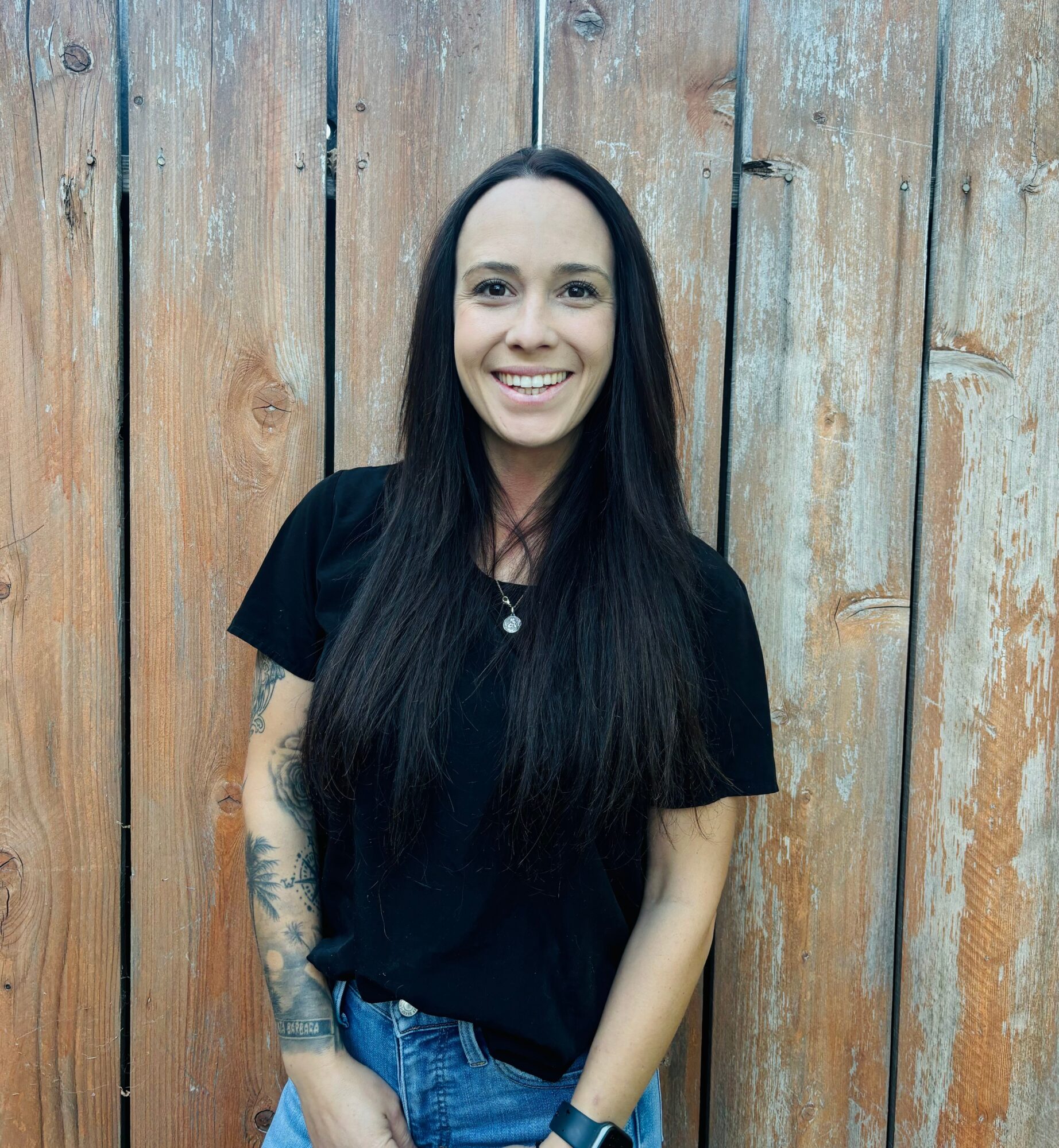 Young woman with long dark hair smiling, standing in front of a wooden fence, wearing a black shirt and jeans.