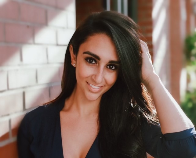Young woman with long dark hair smiling, wearing a dark top, outdoors near a brick wall, sunlight on her face.