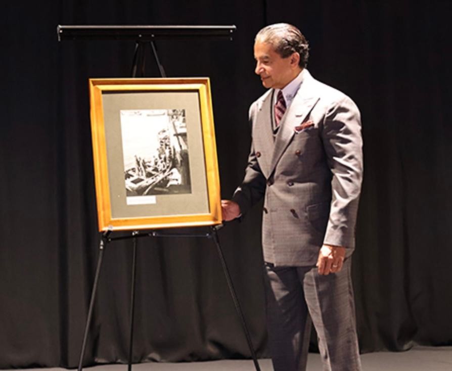 Man in a gray suit looking at framed black and white photograph on stand, black background, stage setting.