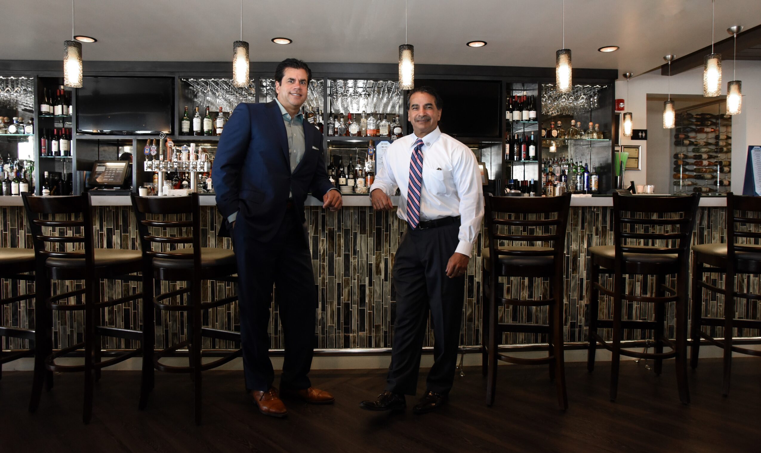 Two men in formal attire standing in front of a bar with high chairs, bottles, and glasses behind them.