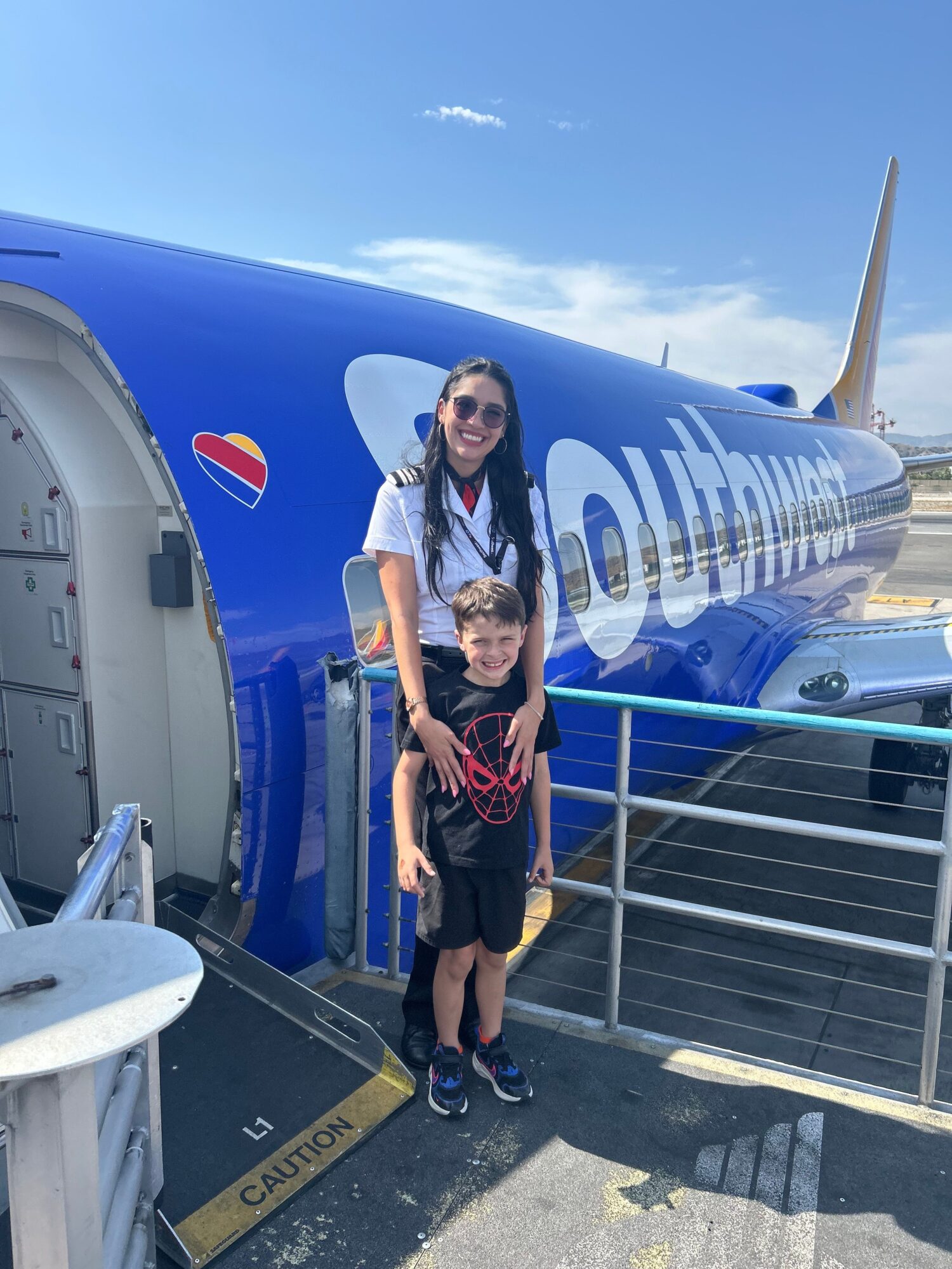 Two women stand in front of an airplane at the gate, smiling, with a clear blue sky in the background.