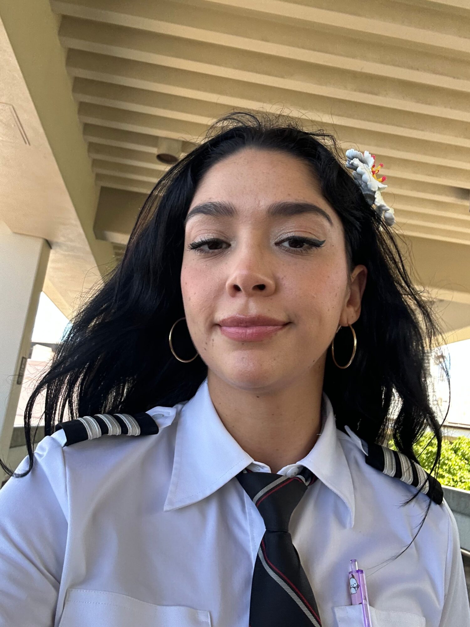 Young woman with dark hair, wearing a white shirt and tie, smiling outdoors near a building with vertical blinds.