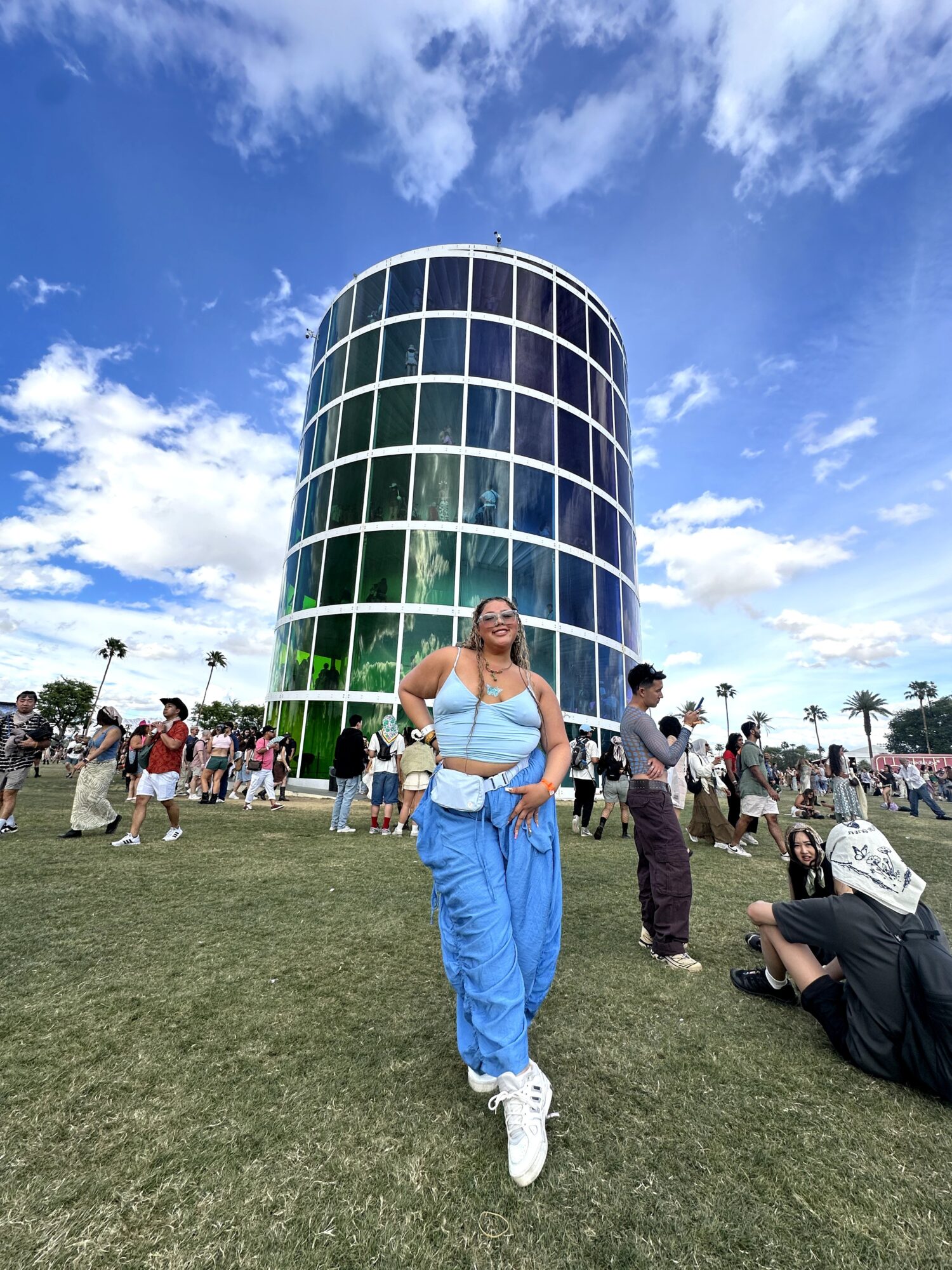 Woman in blue pants and white sneakers standing on grass in front of a tall, cylindrical glass building with a blue sky background.