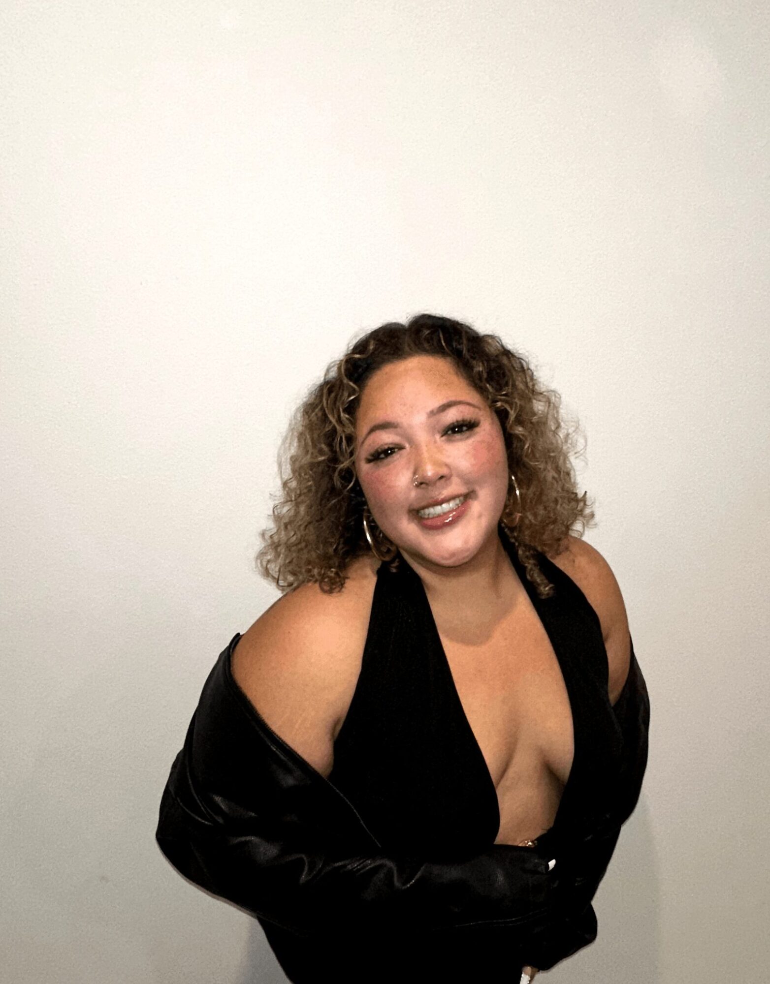 Smiling woman with curly hair wearing a black dress with a deep neckline, standing against a plain white background.