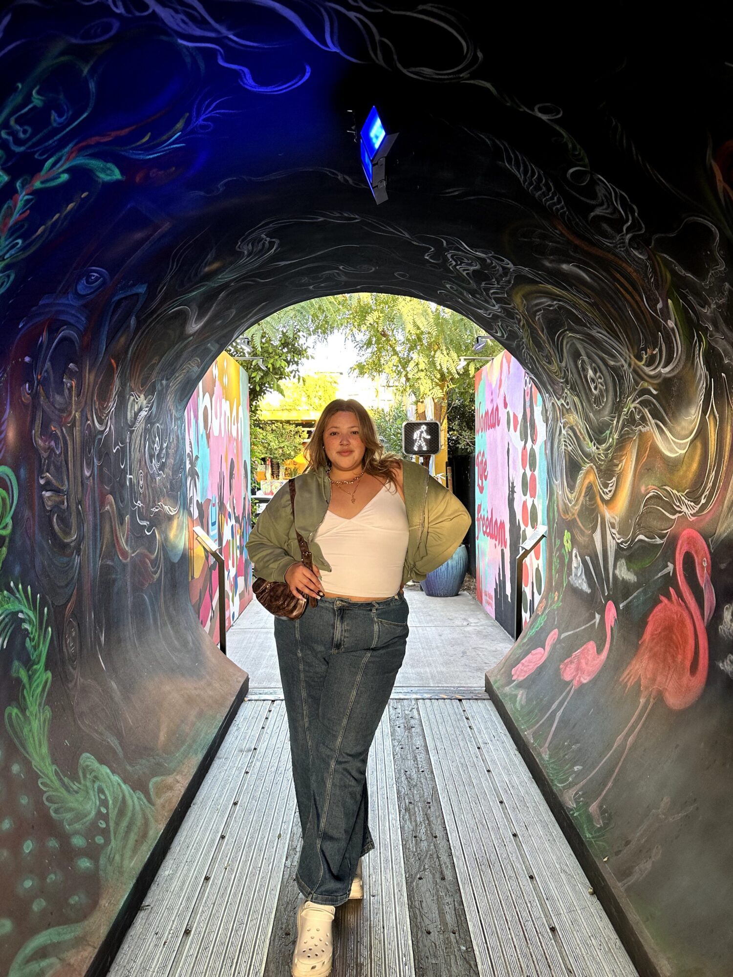 Woman standing in a colorful tunnel with graffiti, trees visible outside, wearing a beige jacket, white top, and jeans.