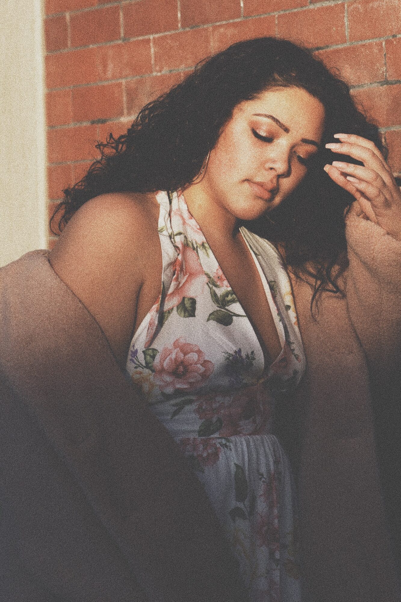 Young woman with dark curly hair, wearing a floral dress, standing against a brick wall, looking down with her hand in her hair.