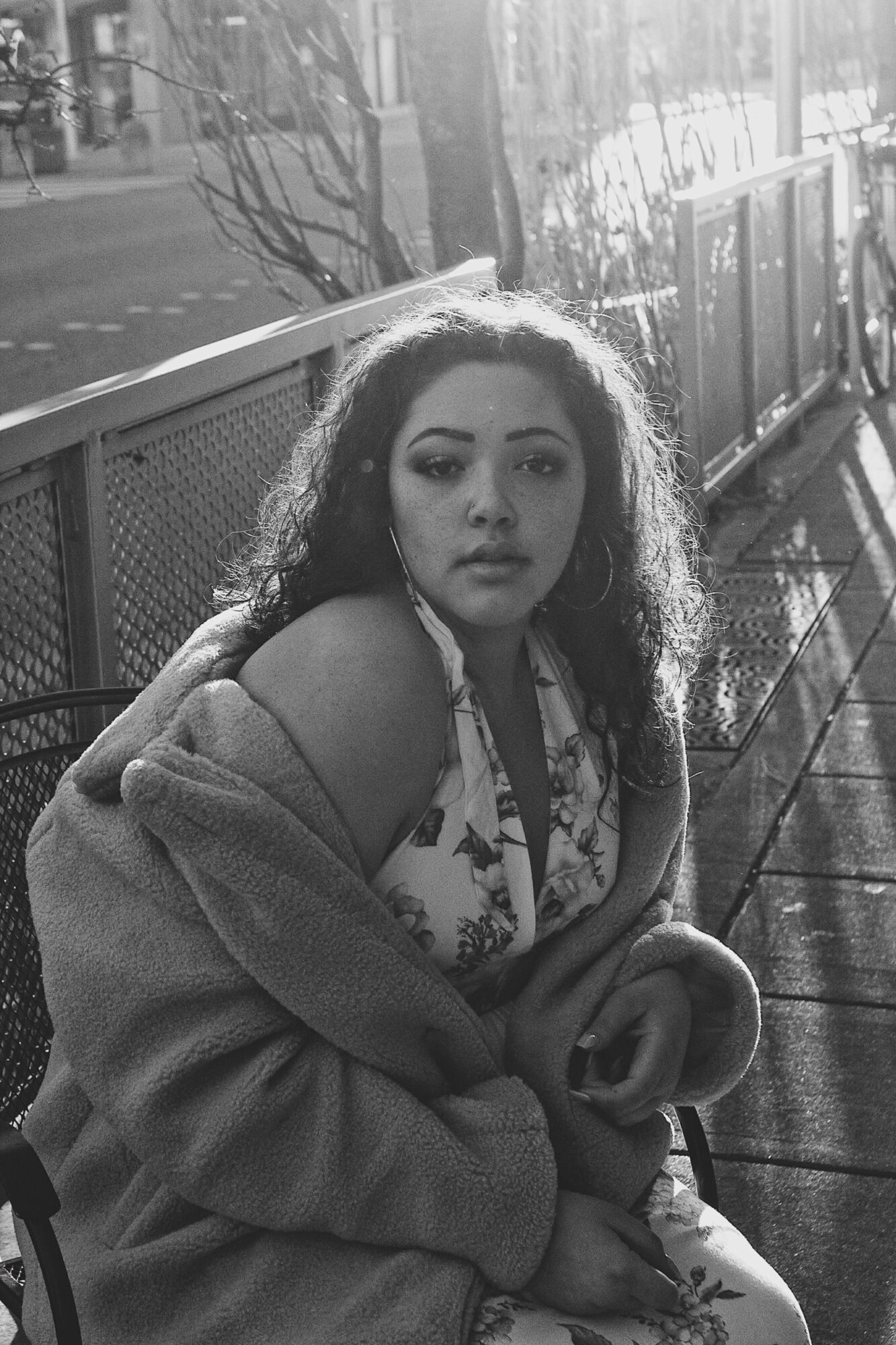 Young woman with curly hair sitting on a bench outdoors, sunlight shining on her face, wearing a coat and patterned dress.
