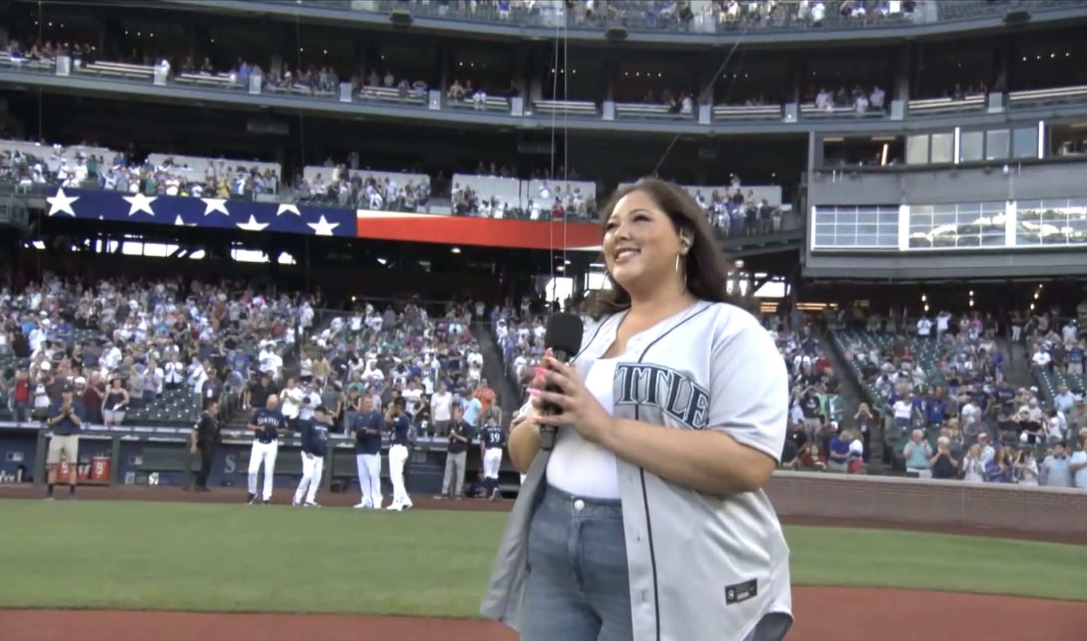 Person smiling and holding a microphone at a baseball stadium with a crowd in the background.
