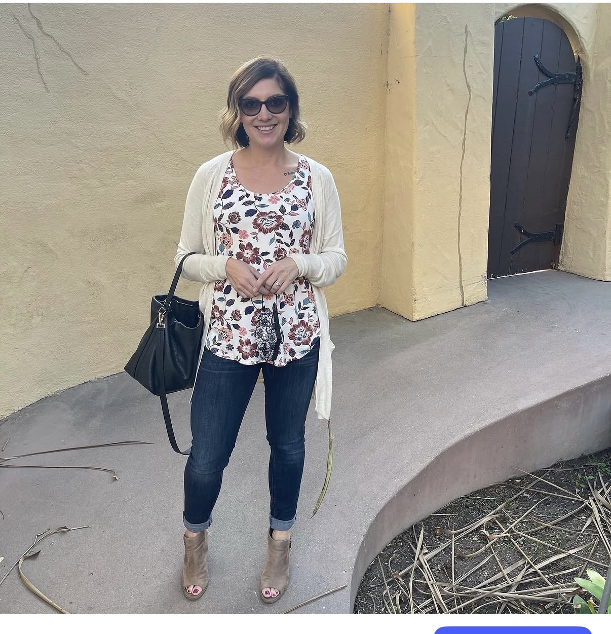 Woman with sunglasses, smiling, standing outdoors against a yellow wall, wearing a floral top, jeans, and beige heels, holding a black bag.