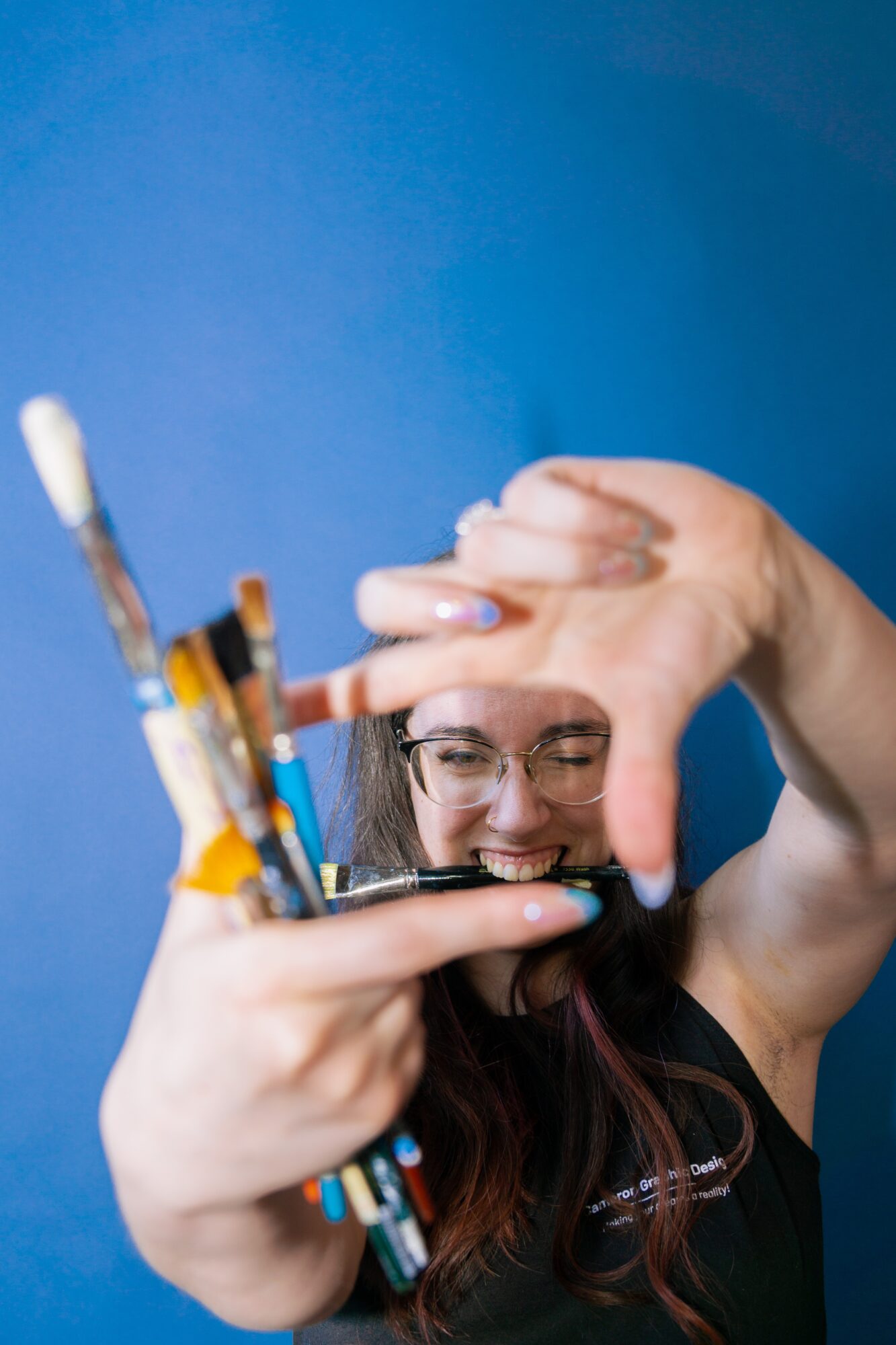 Person with glasses smiling, holding paintbrushes and a pen, against a blue background.