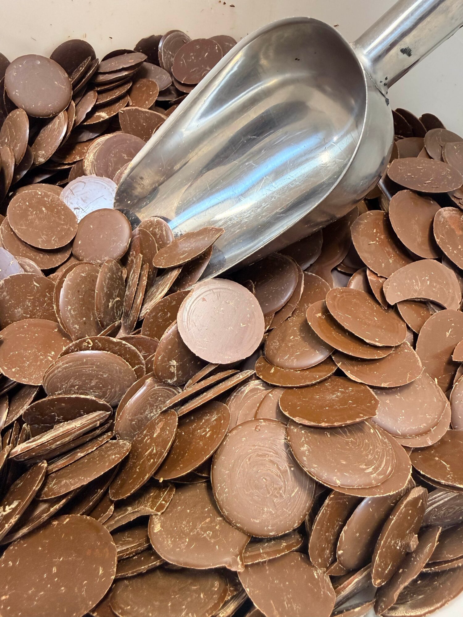 A metal scoop partially submerged in a pile of round chocolate candies, some spilling out of the scoop.