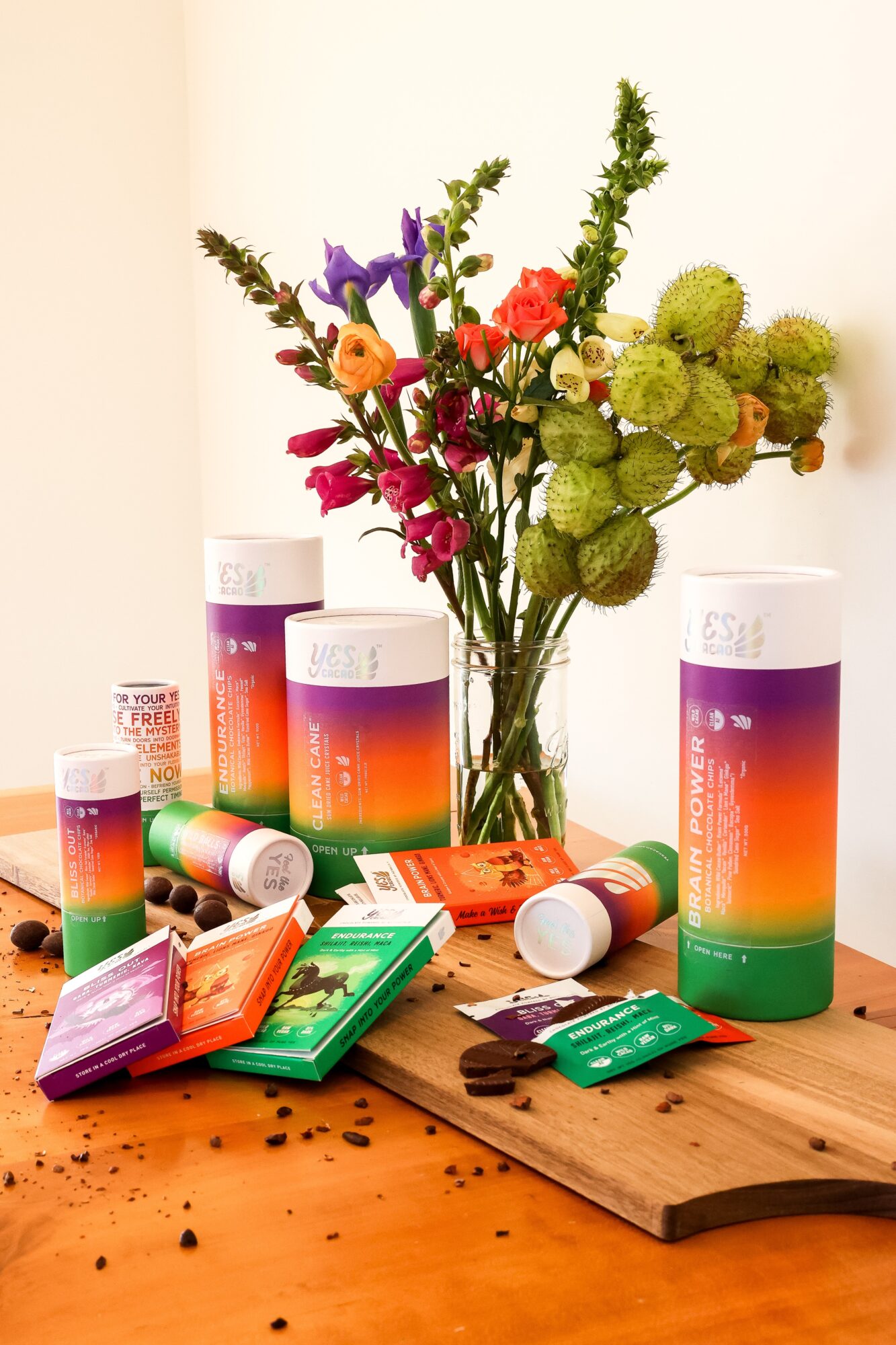 Colorful herbal tea packages and a bouquet of flowers on a wooden surface, with a plain background.