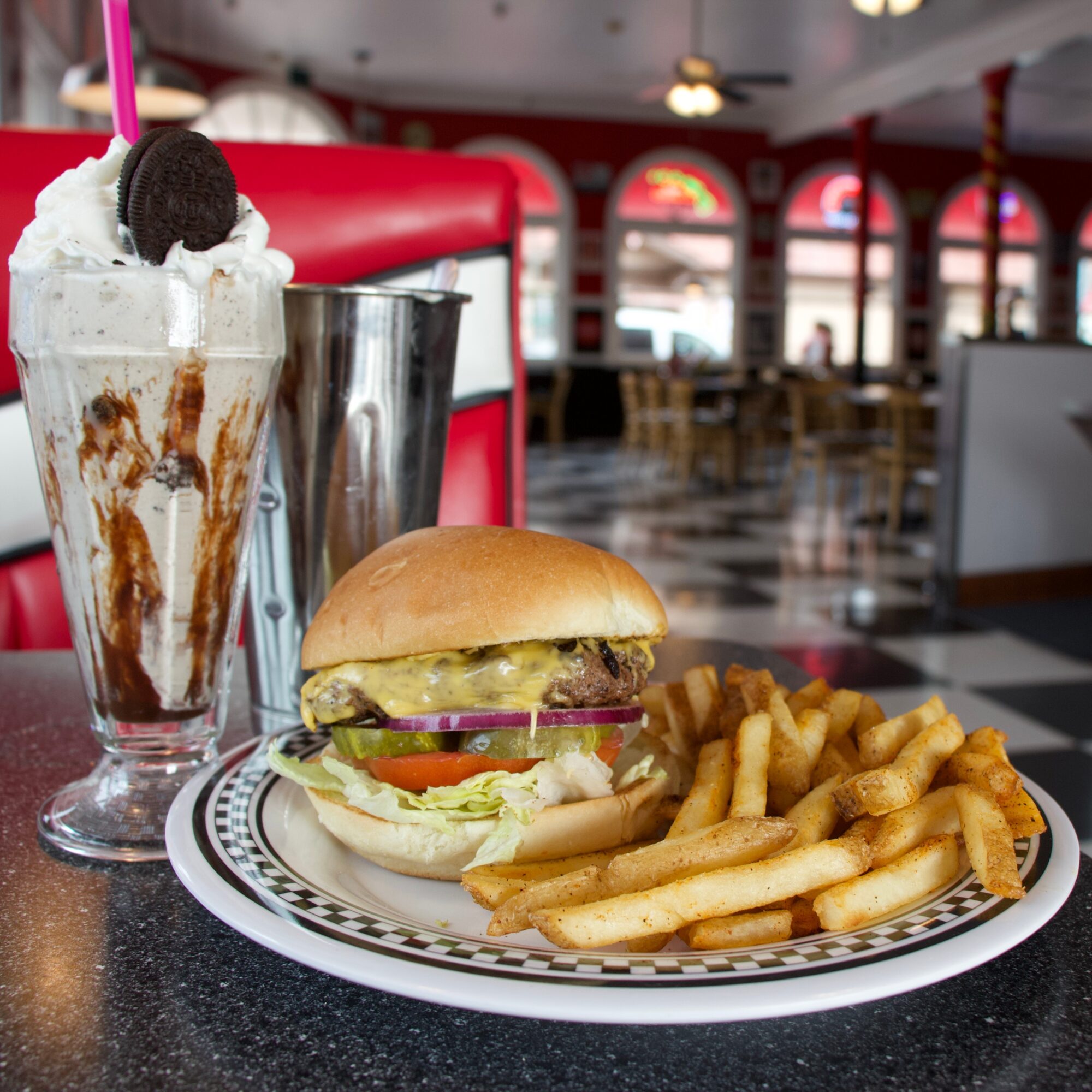 Milkshake with whipped cream, chocolate syrup, and an Oreo cookie, burger with lettuce, tomato, pickles, and fries on a plate, in a diner setting