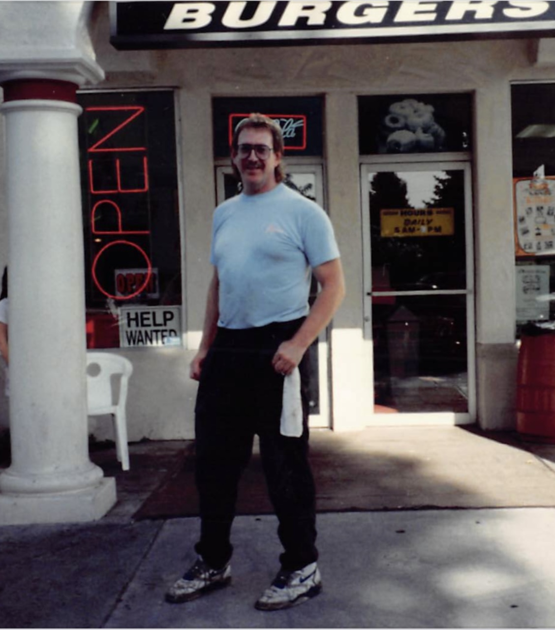 Man standing outside a burger restaurant, smiling, wearing a light blue shirt and dark pants, with a white towel in hand.