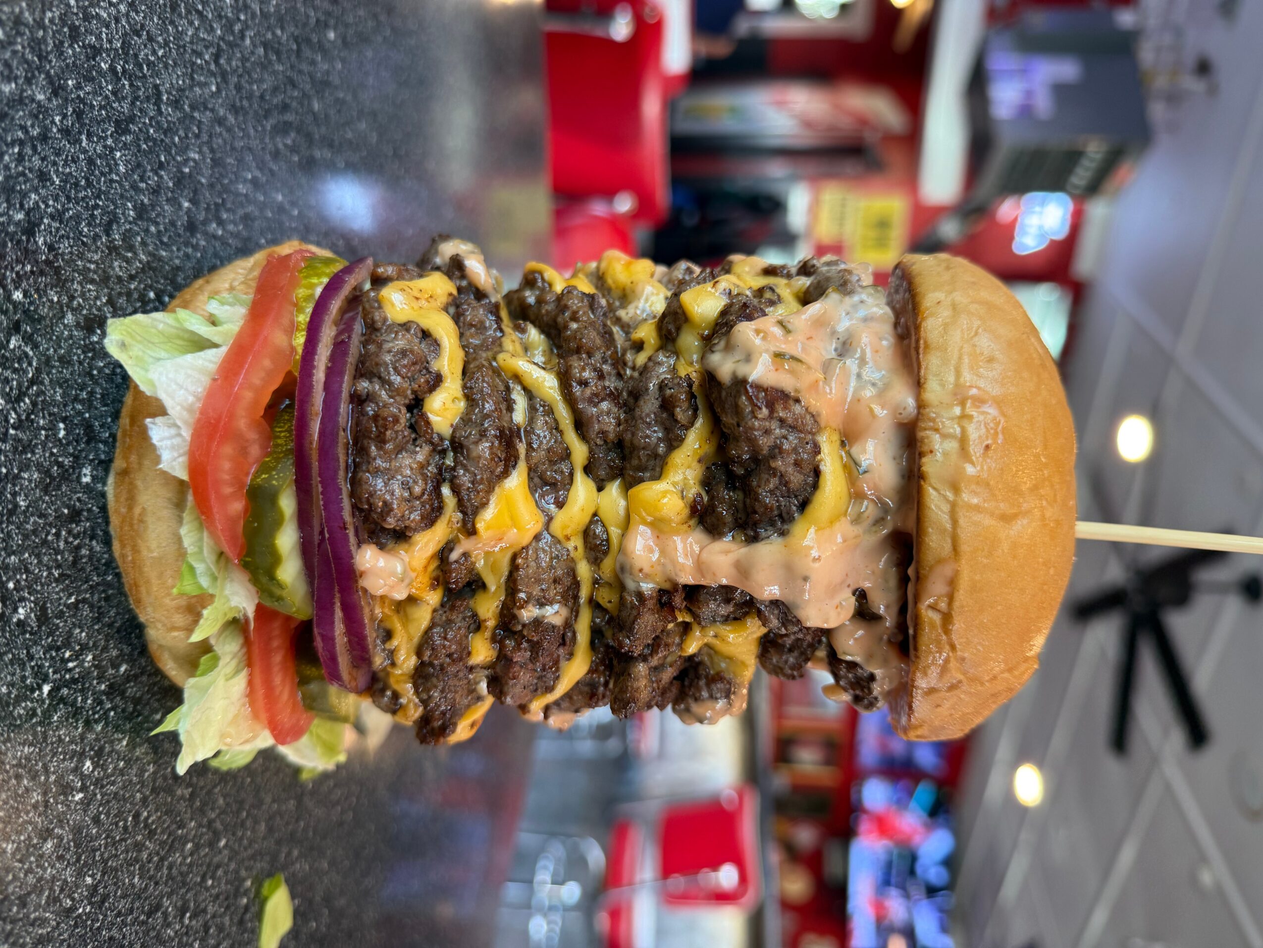 A burger with lettuce, tomato, red onion, cheese, and multiple beef patties stacked in a bun, on a black surface.