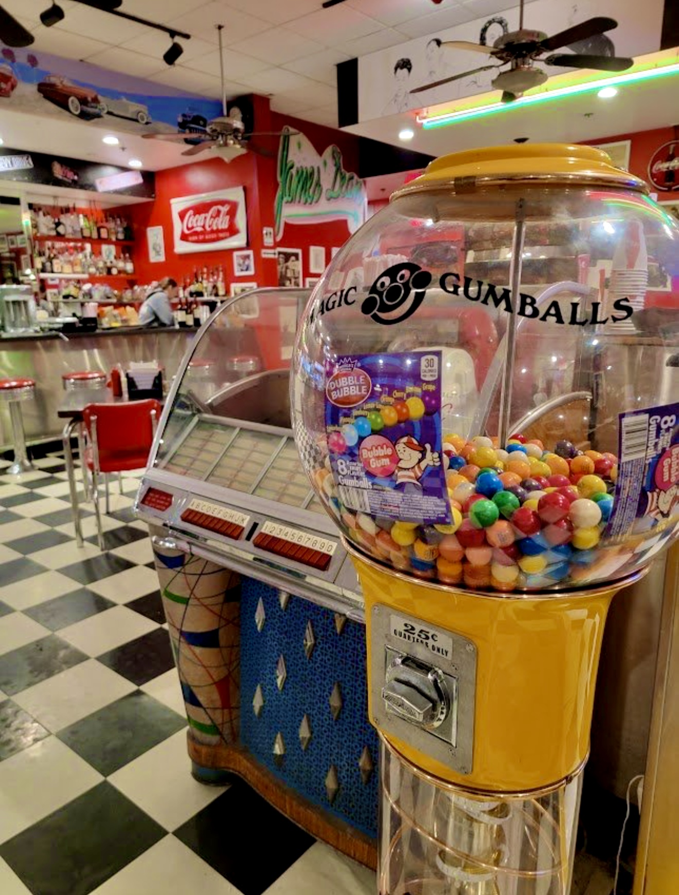 Gumball machine filled with colorful gumballs in a retro-style arcade or diner setting, checkered floor, red walls, and ceiling fans.