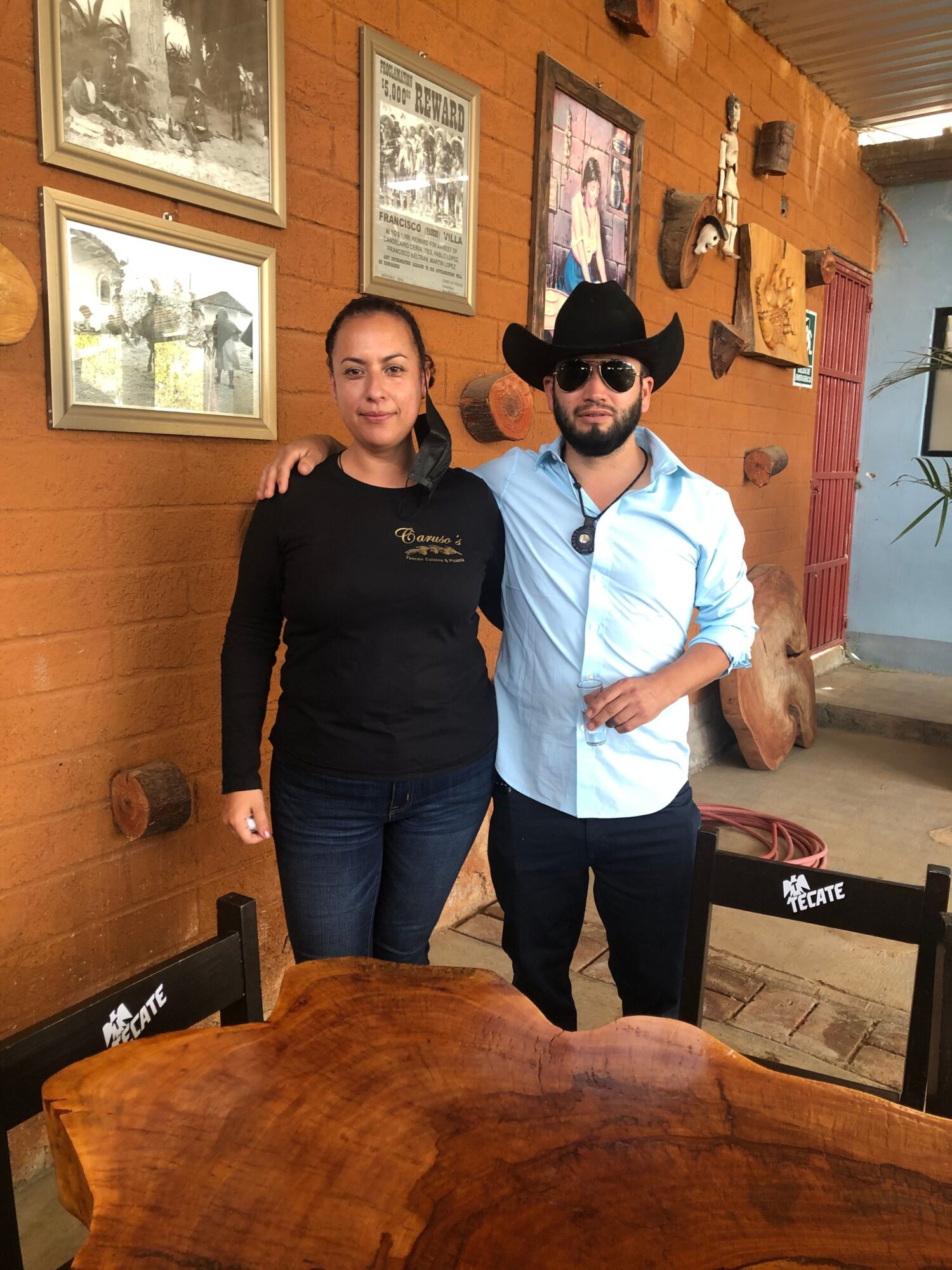 Two women standing indoors, one wearing sunglasses and a light blue shirt, the other in a black jacket, wooden walls, framed pictures, and a wooden table.