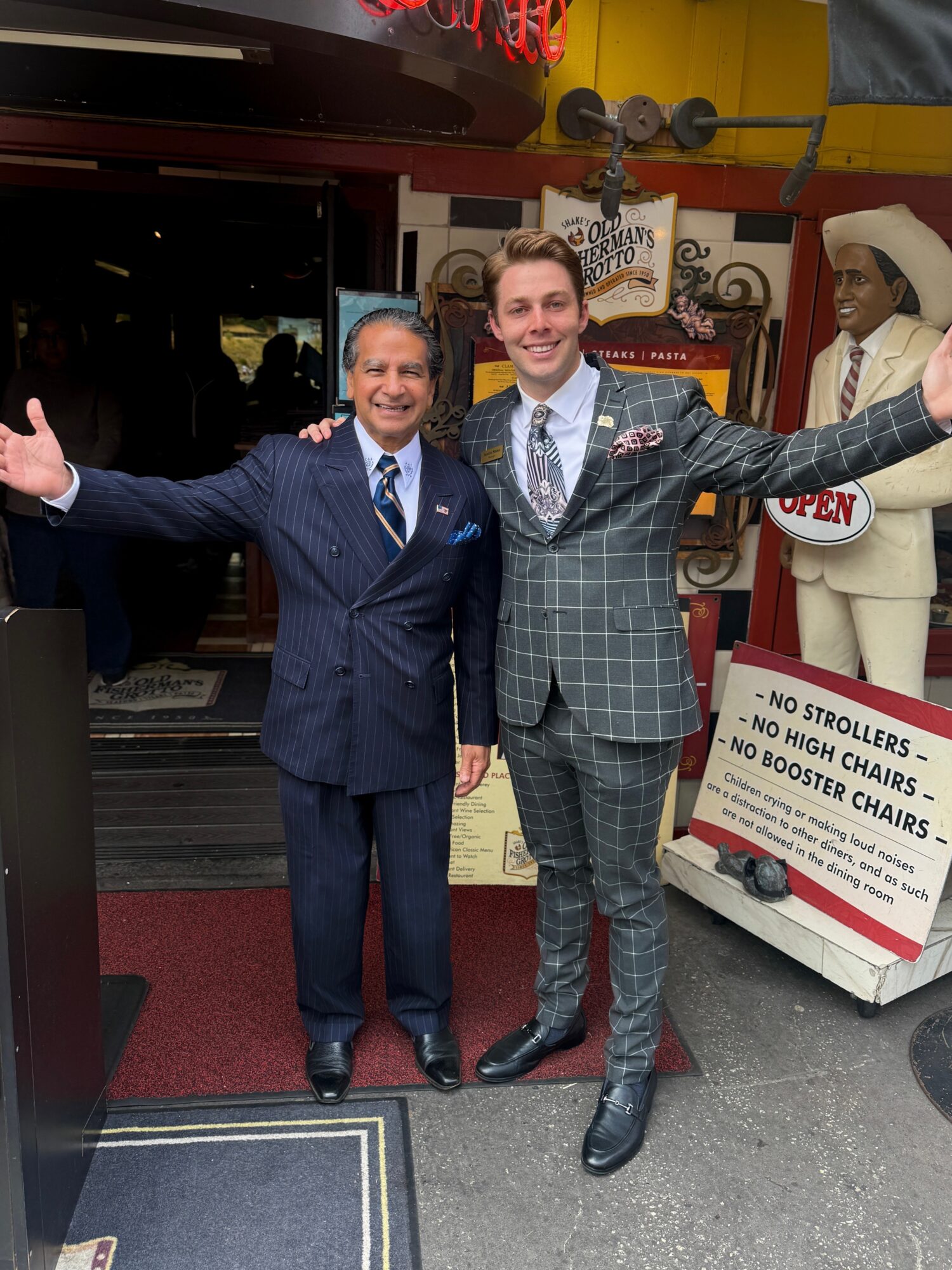 Two men in suits stand outside a venue, smiling with arms outstretched. A mannequin in a white suit and hat is nearby. Word signs are visible.
