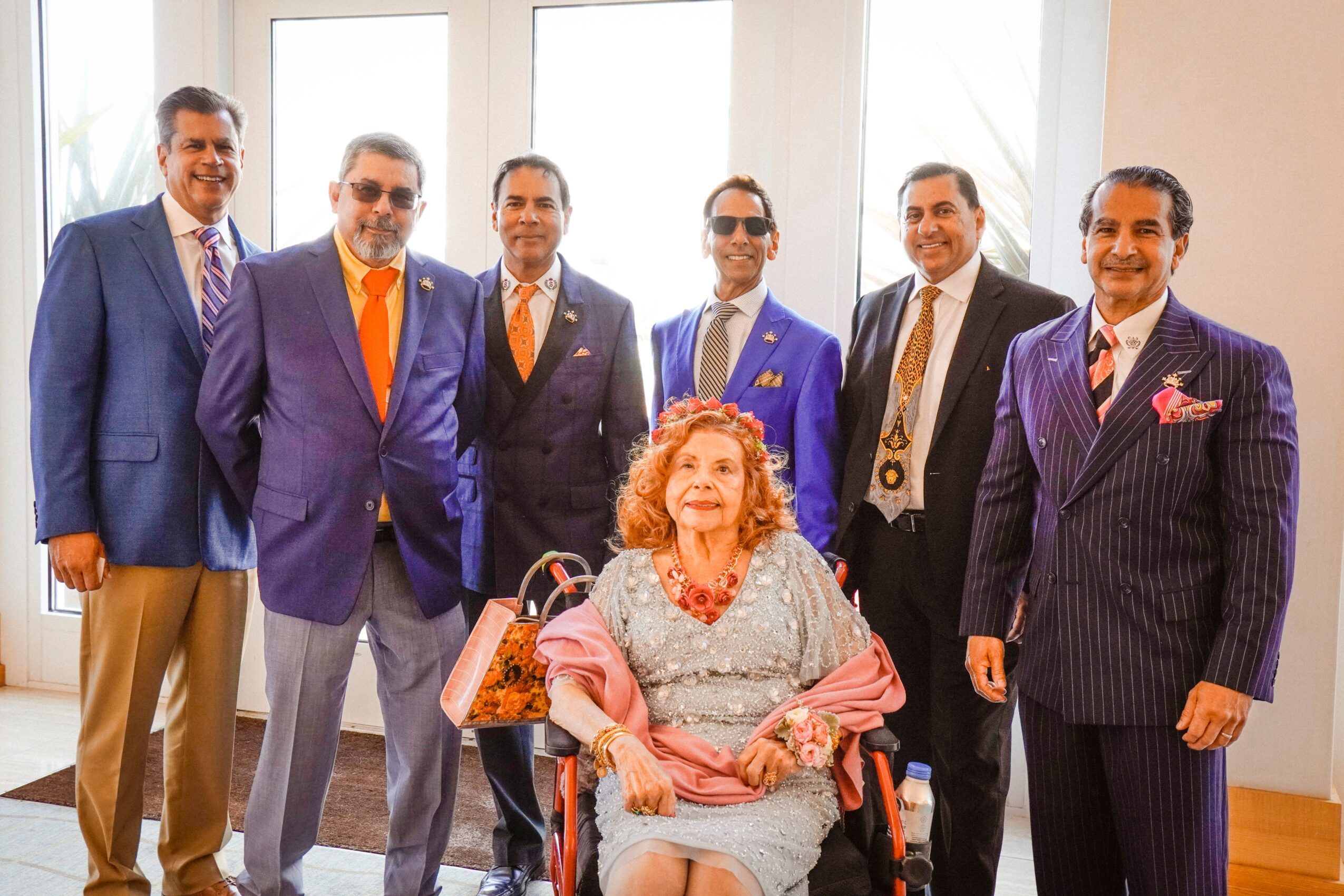 Six men in suits standing behind a seated woman with curly hair in a bright room with large windows.