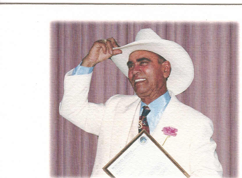 Man in white suit and hat smiling, holding a framed certificate, with a pink flower on his lapel, against a purple curtain background.