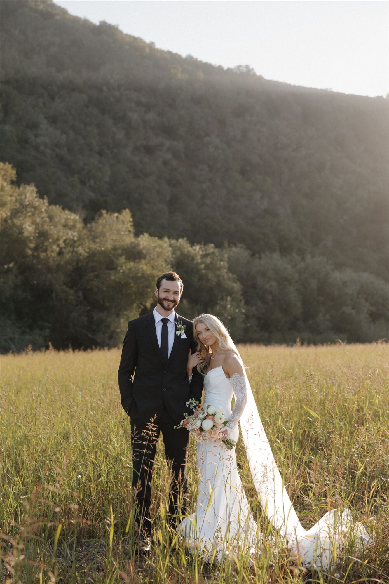 A bride and groom stand in a grassy field with a hillside and trees in the background, sunlight shining behind them.