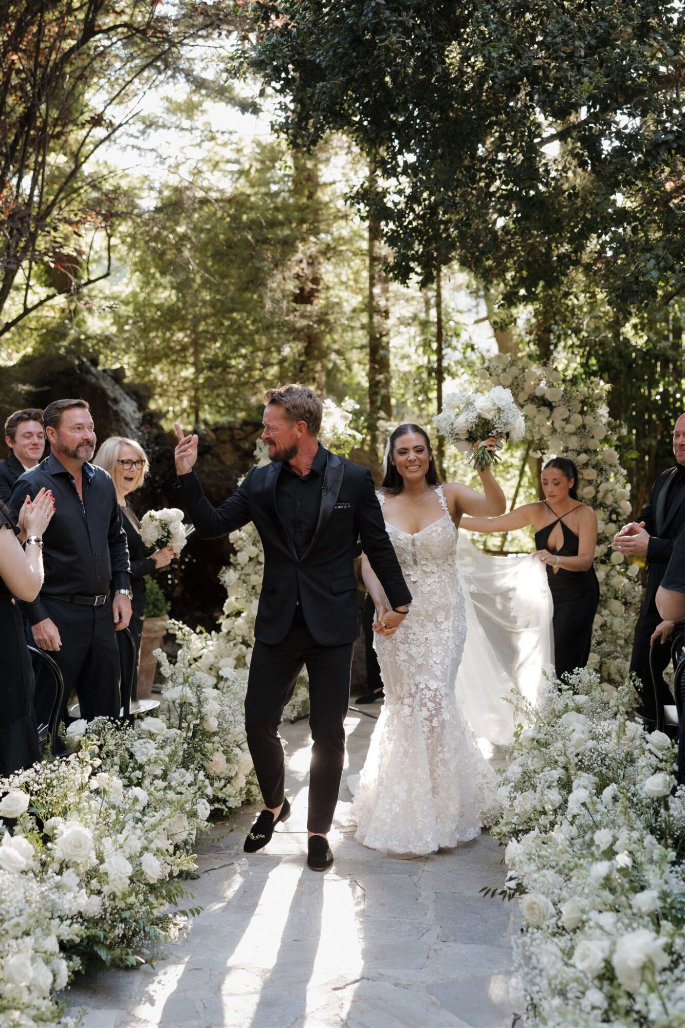 Couple walking down an outdoor aisle with guests on both sides, surrounded by white flowers and trees, during a wedding ceremony.