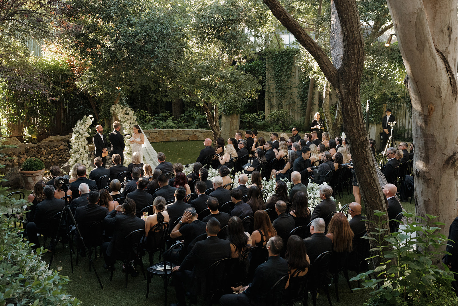 Wedding ceremony outdoors with guests seated, bride and groom standing, surrounded by trees and greenery.