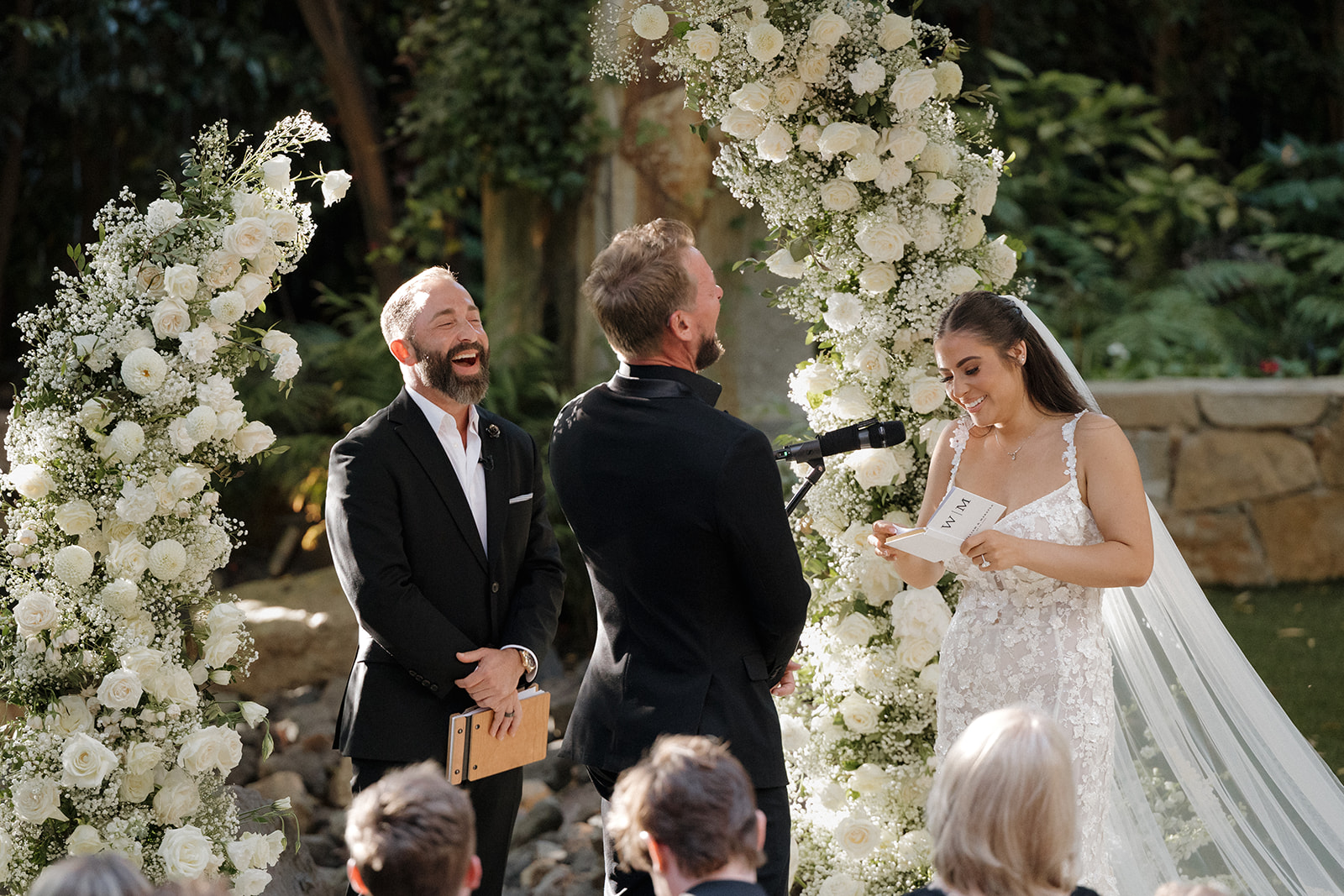 Bride and groom exchanging vows outdoors, surrounded by floral arches, with guests watching, in a garden setting.