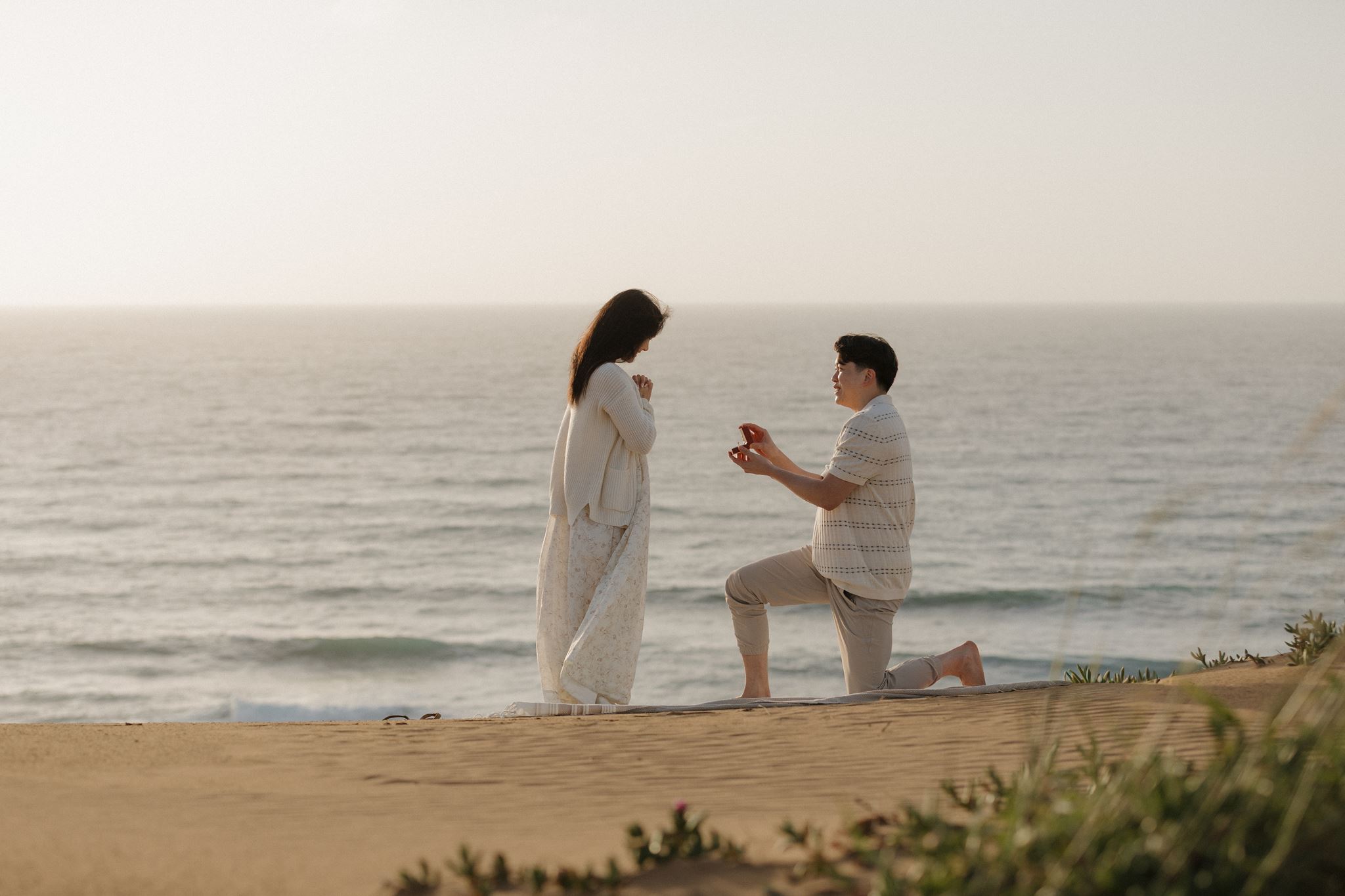 A man kneeling on one knee proposes to a woman on a beach, with the ocean in the background.