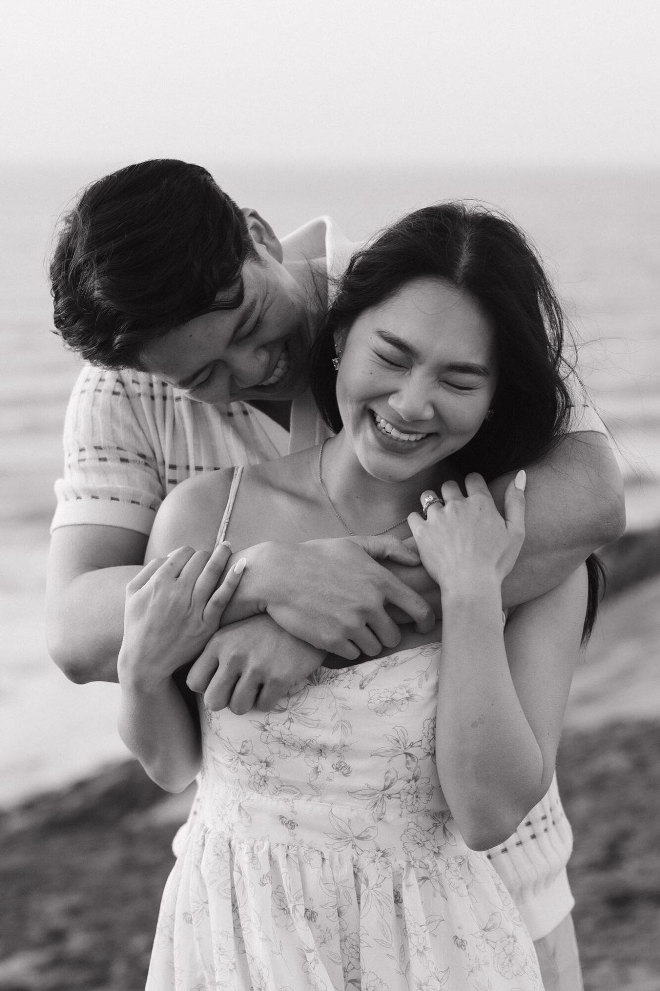 Two people smiling and hugging on a beach, one with arms around the other, in black and white.