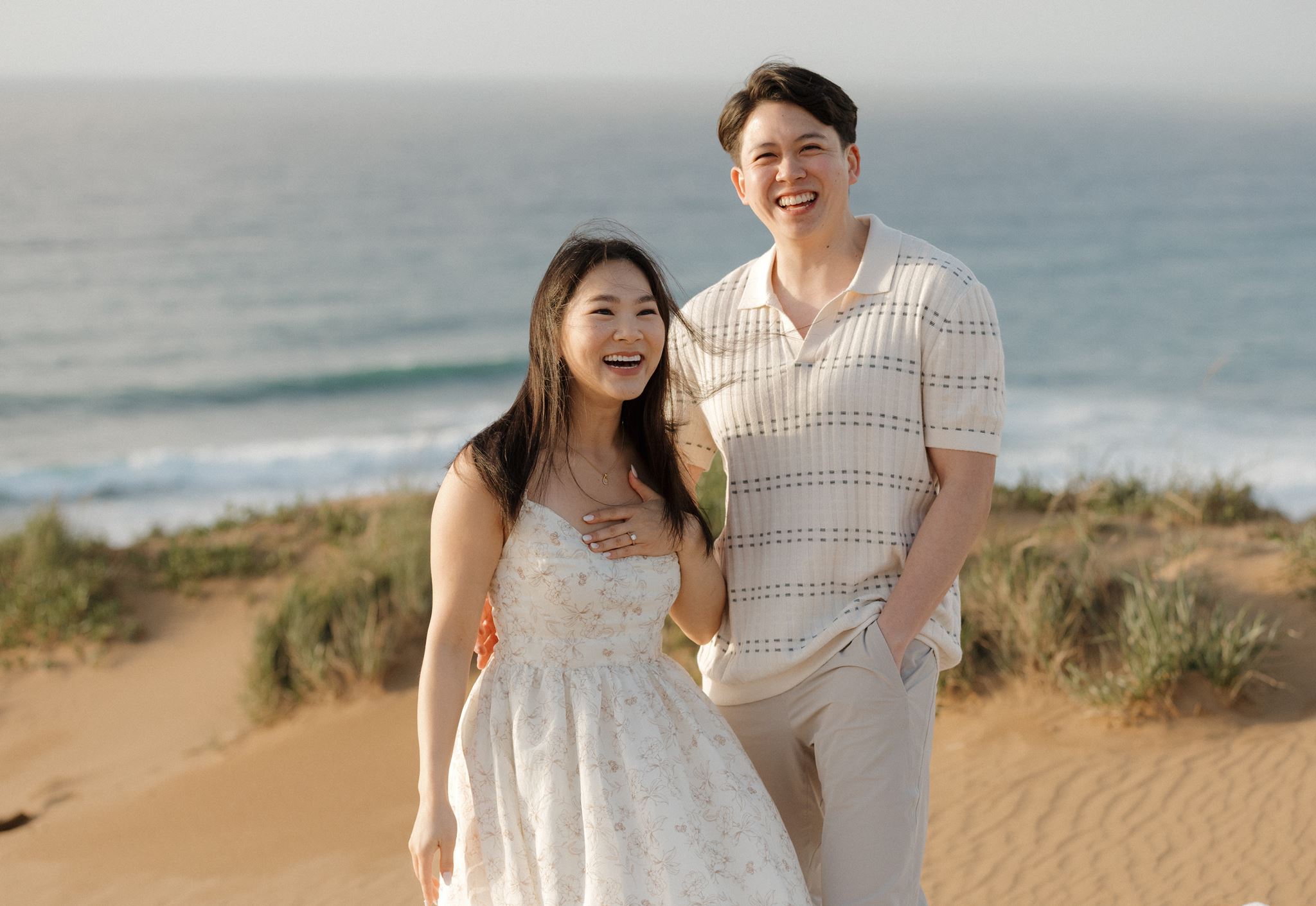 A smiling couple standing on a sandy beach with ocean waves in the background.