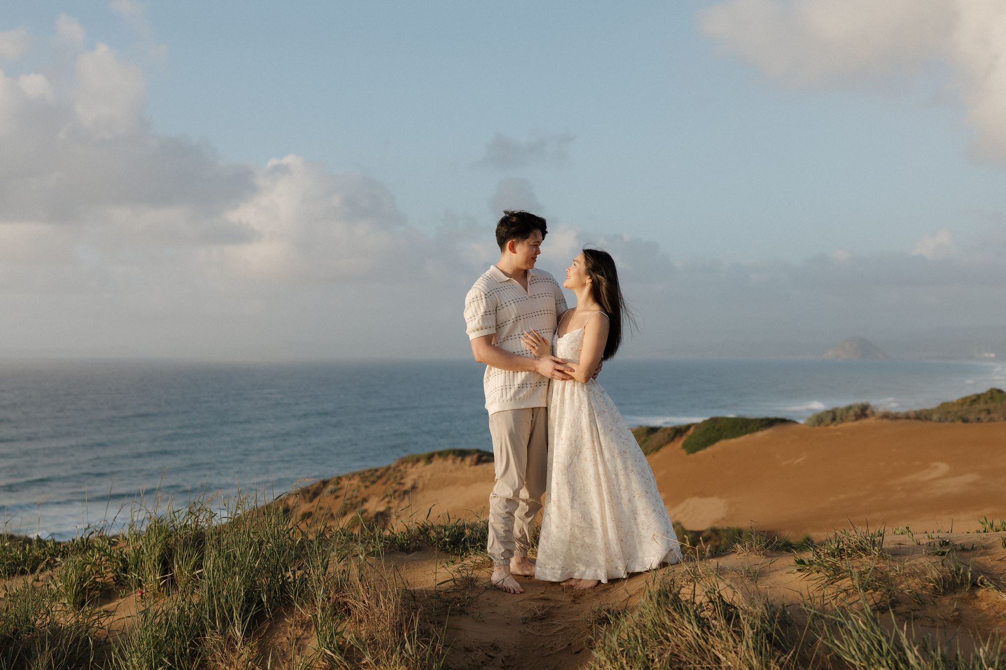 A couple stands on a grassy hill near the ocean, holding hands and looking at each other, with clouds in the sky.
