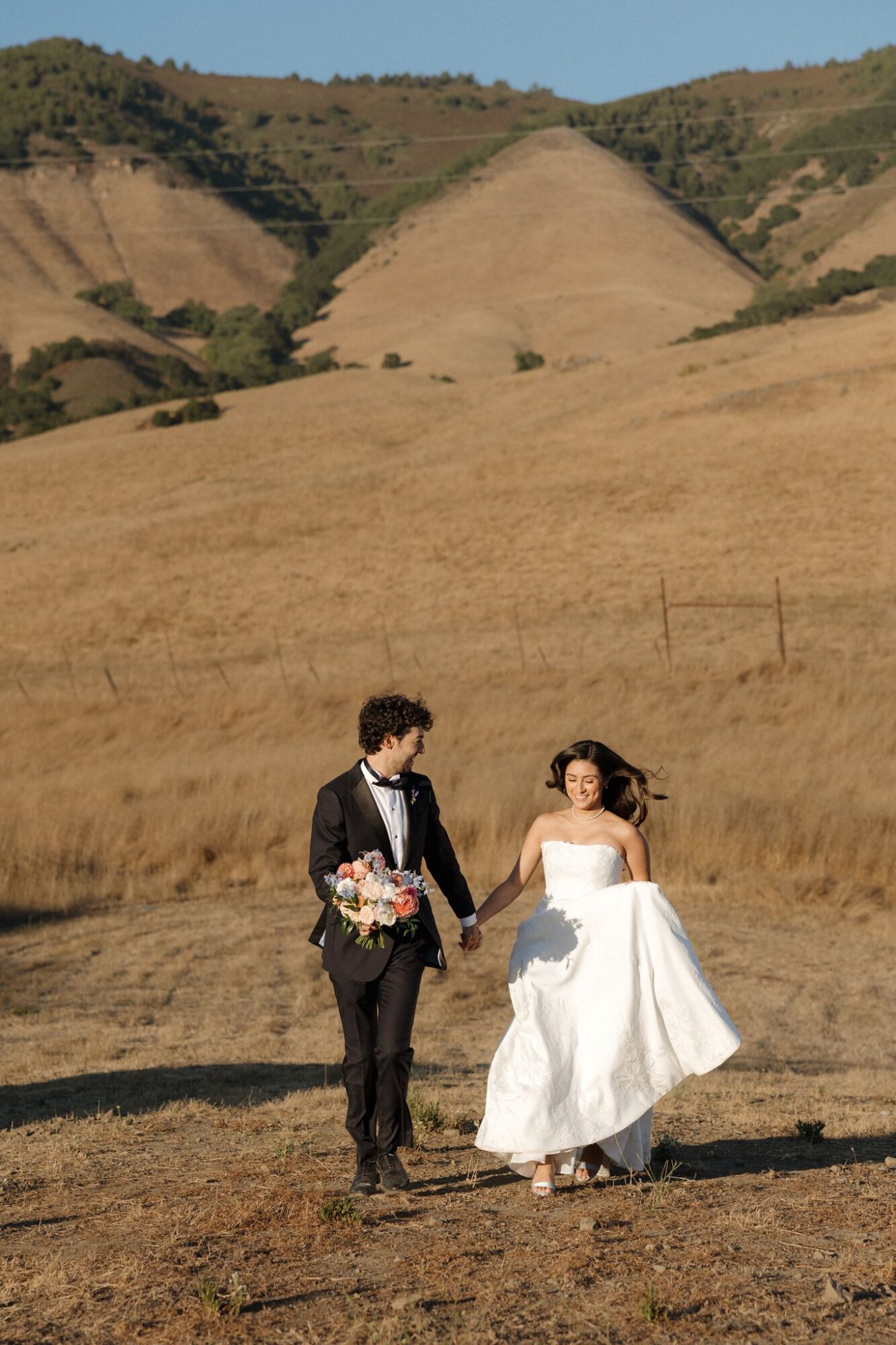 A couple in wedding attire walking hand in hand in a golden field with rolling hills in the background.