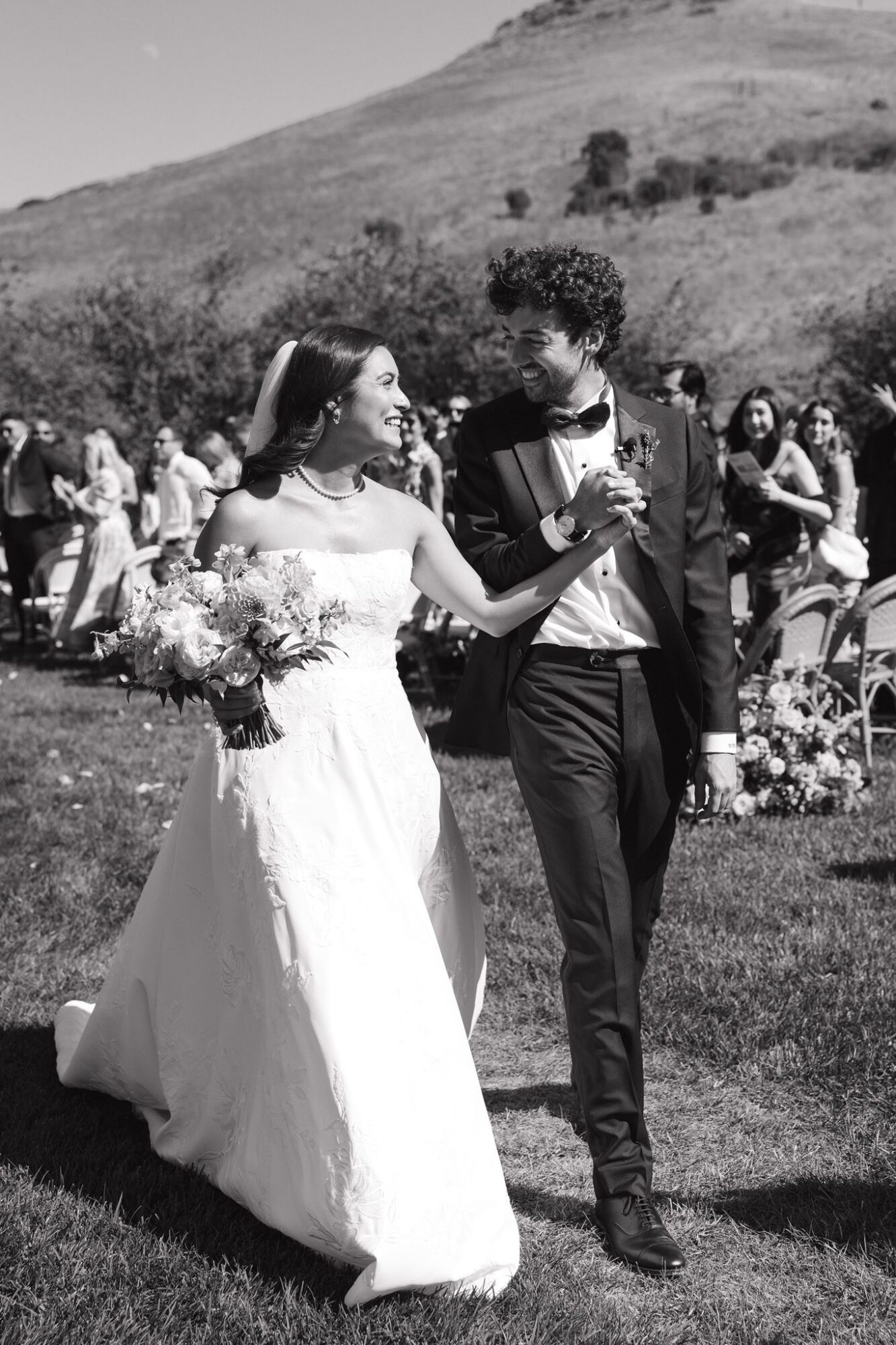 Bride and groom walking outdoors, holding hands, with guests and a hilly landscape in the background.