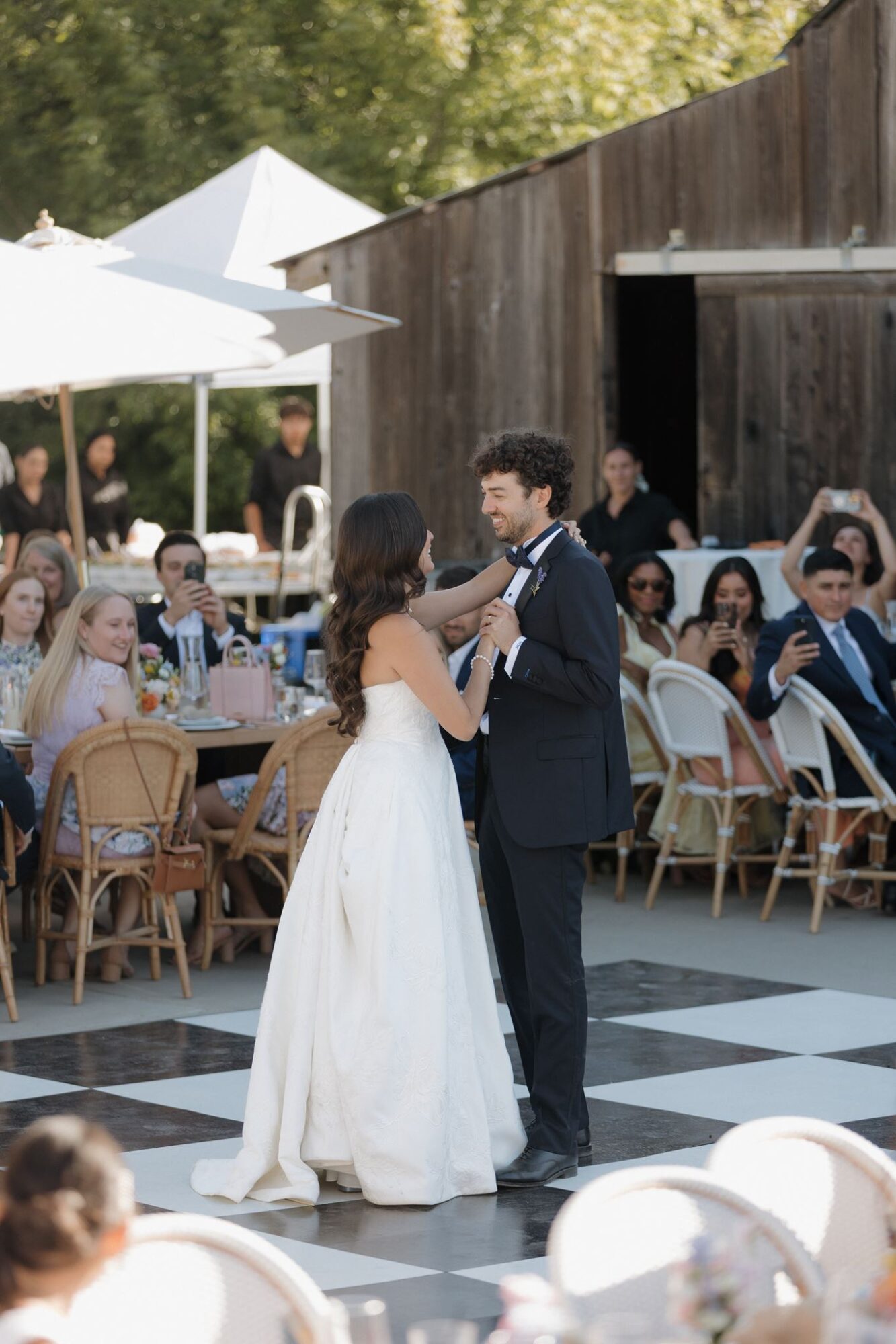 Couple dancing outdoors at wedding reception with seated guests and rustic wooden building in background.