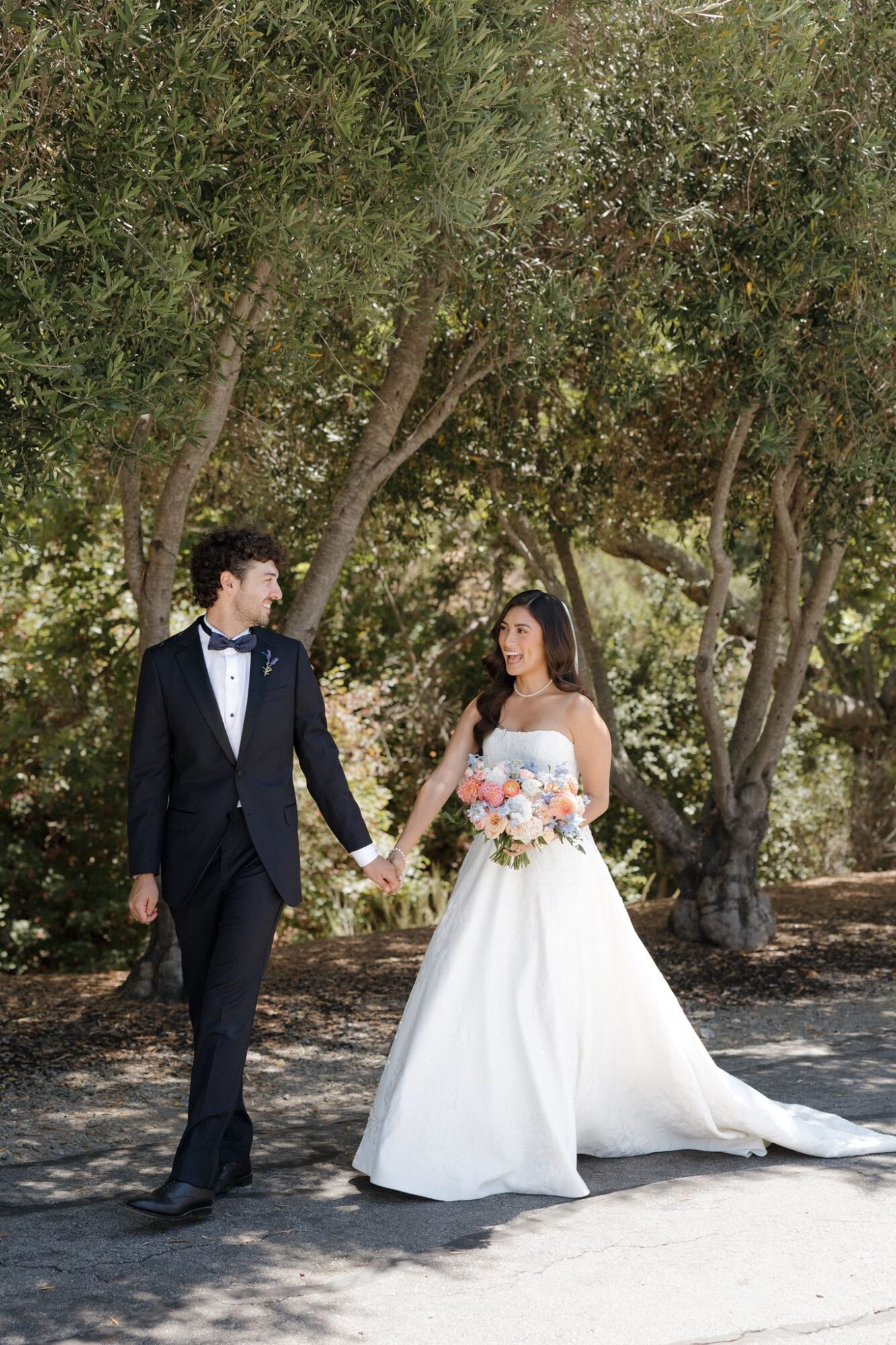 A bride and groom holding hands outdoors, surrounded by trees, with the bride in a white gown and the groom in a tuxedo.