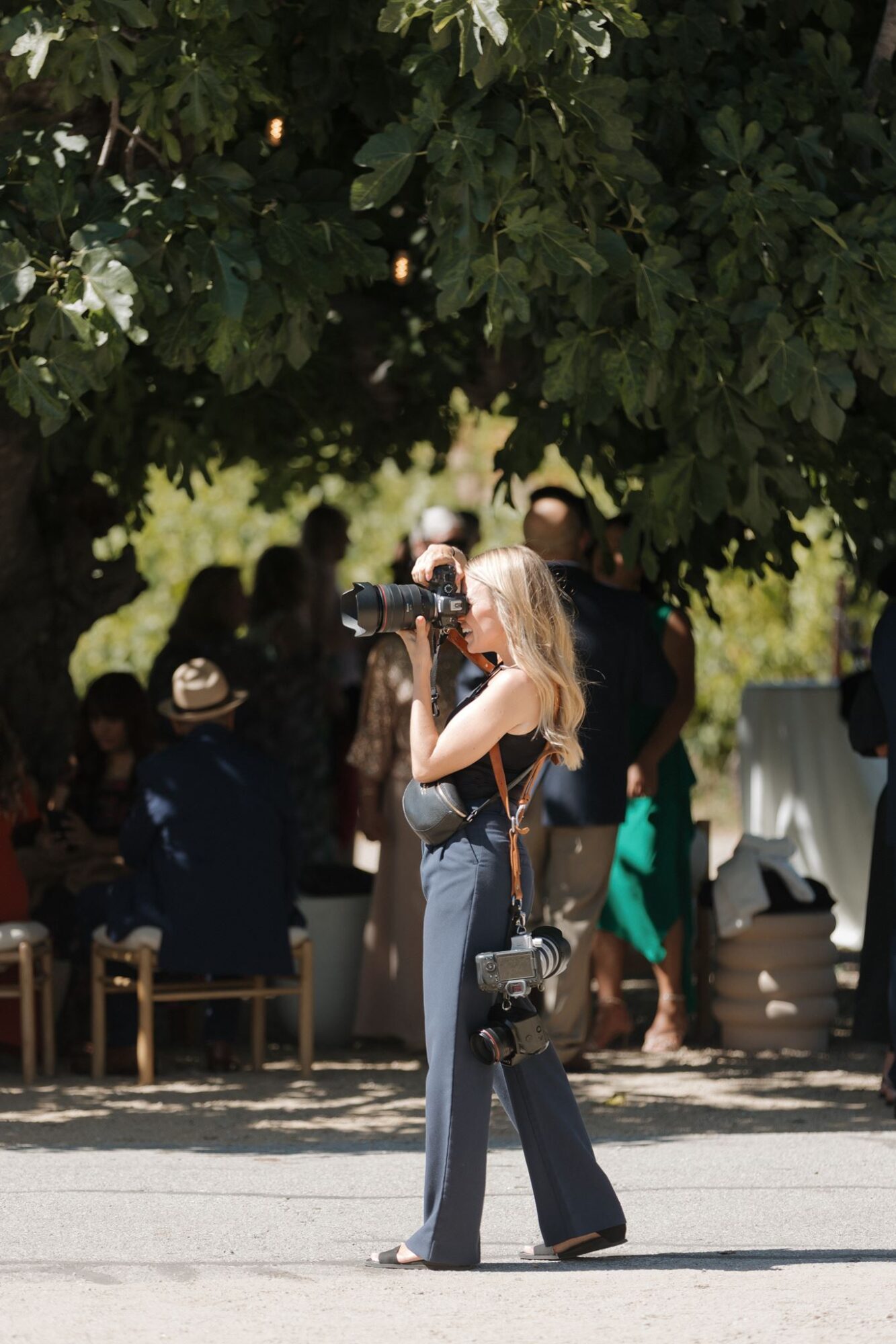 Woman with long blonde hair taking a photo outdoors under a large leafy tree, with people sitting and standing in the background.