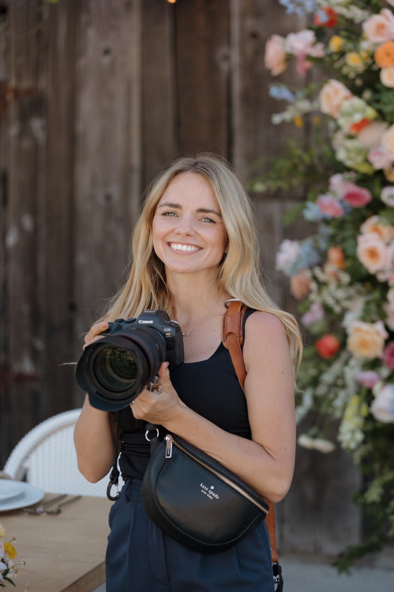 Young woman with blonde hair smiling, holding a camera, standing outdoors near a wooden fence and colorful flowers.