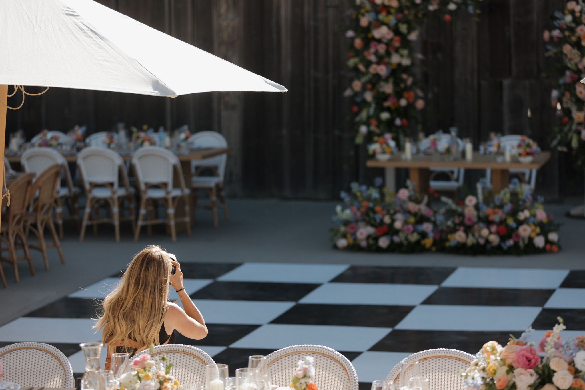 A woman with long hair sitting at a table outdoors, with a checkered dance floor and floral decorations in the background.