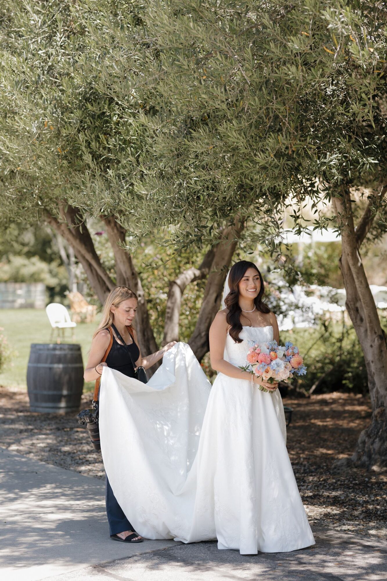 Two women outdoors, one holding a bouquet, surrounded by trees and sunlight, with a grassy area in the background.