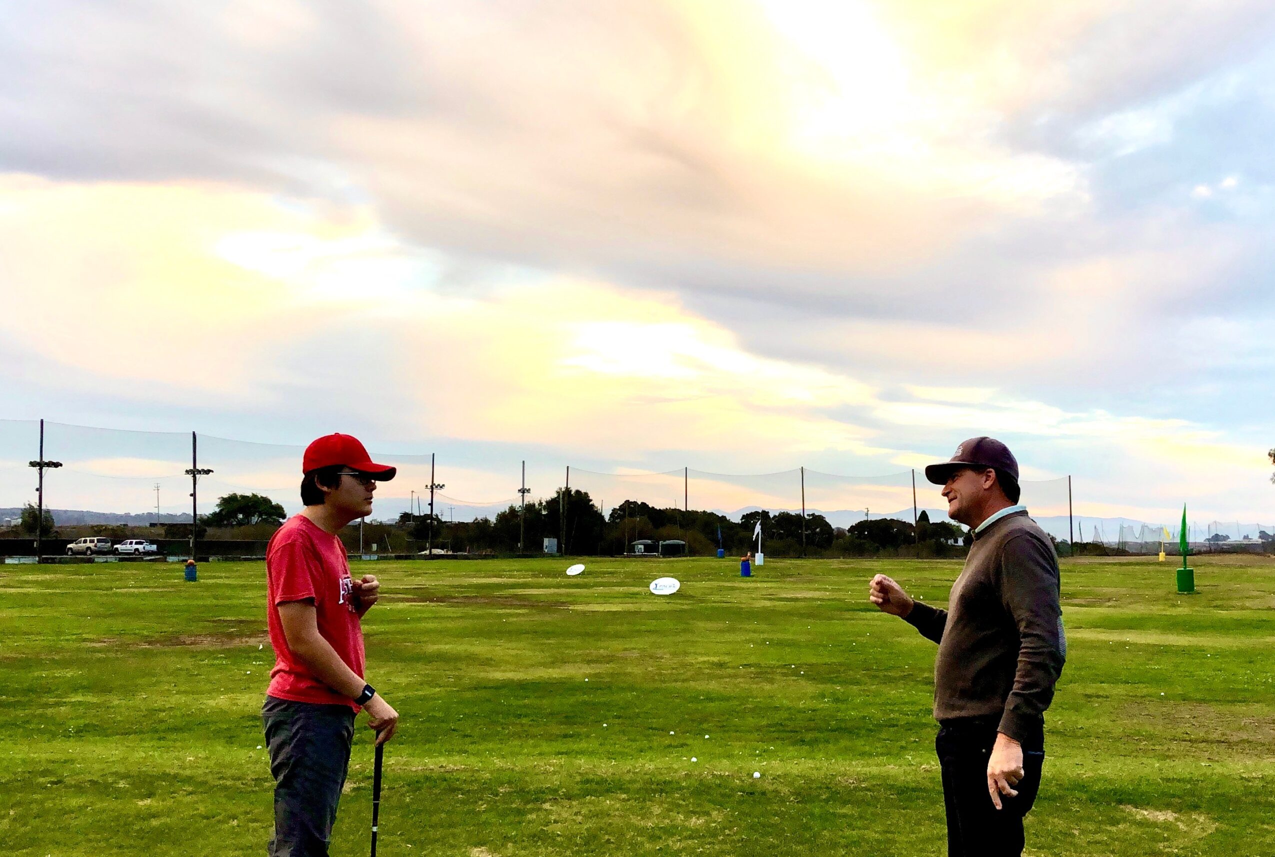 Two people standing on a golf course, one holding a golf club, engaging in conversation under a cloudy sky.