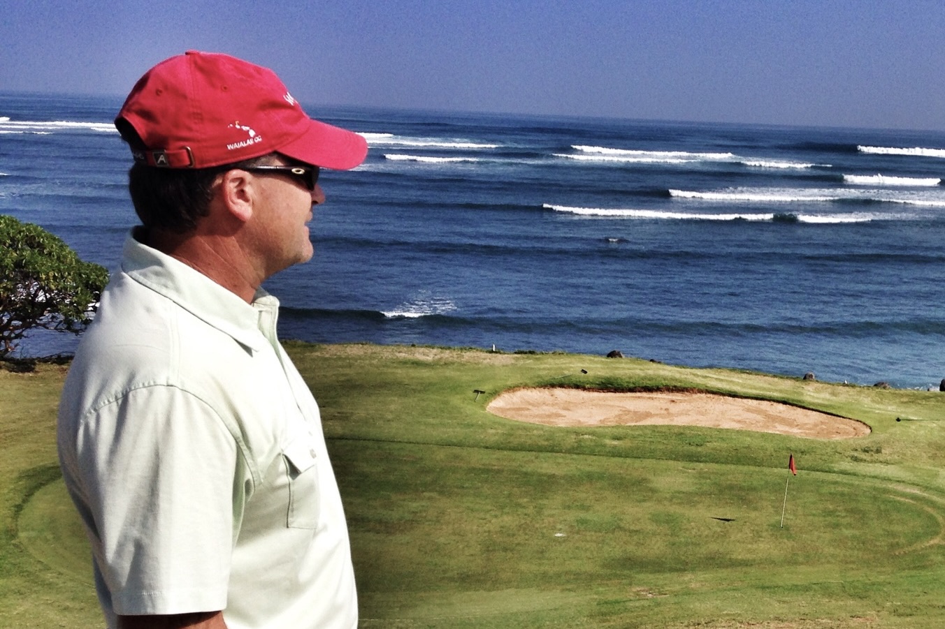 Man in white shirt and red cap standing on a golf course near the ocean, with waves in the background.