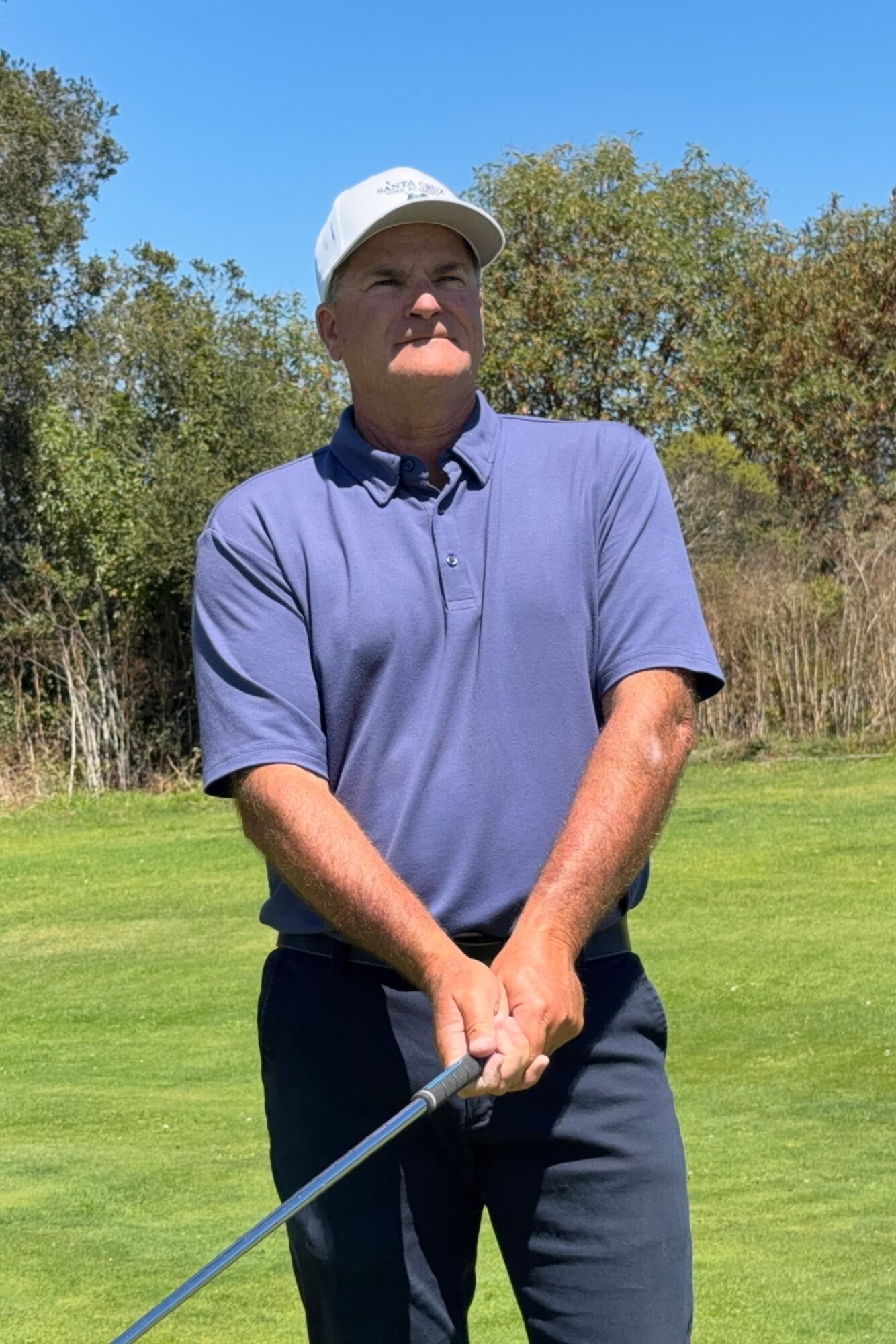 Man in a blue polo shirt and white cap holding a golf club outdoors, with trees and blue sky in background.
