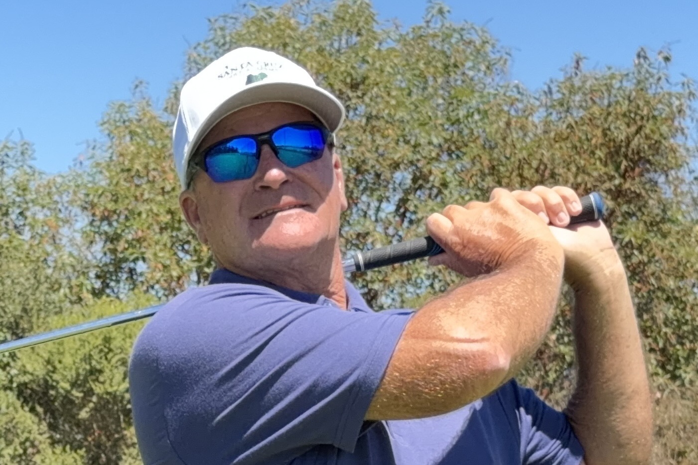 Man wearing sunglasses, white cap, and blue shirt holding a golf club outdoors with trees in background.