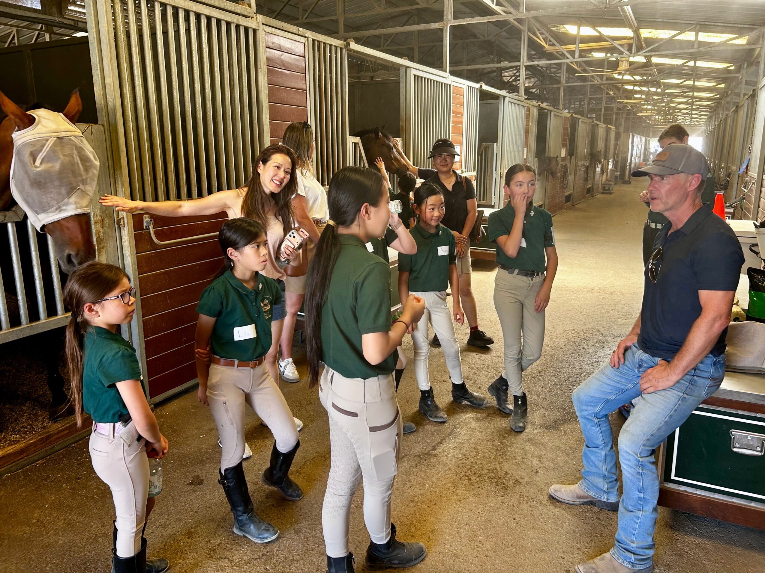 Group of children in green shirts and beige pants gathered around a man sitting on a bench inside a stable or barn.